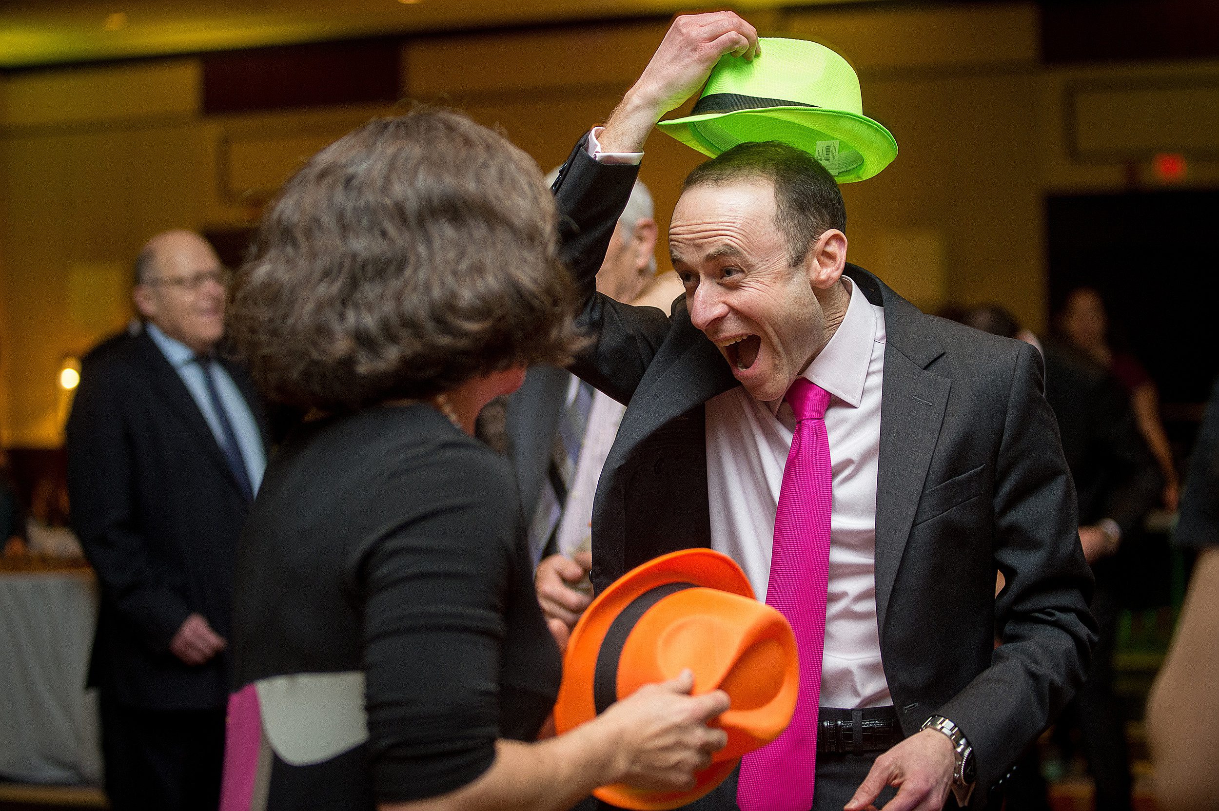 A man celebrates and plays in a black suit and a neon green fedora and pink tie