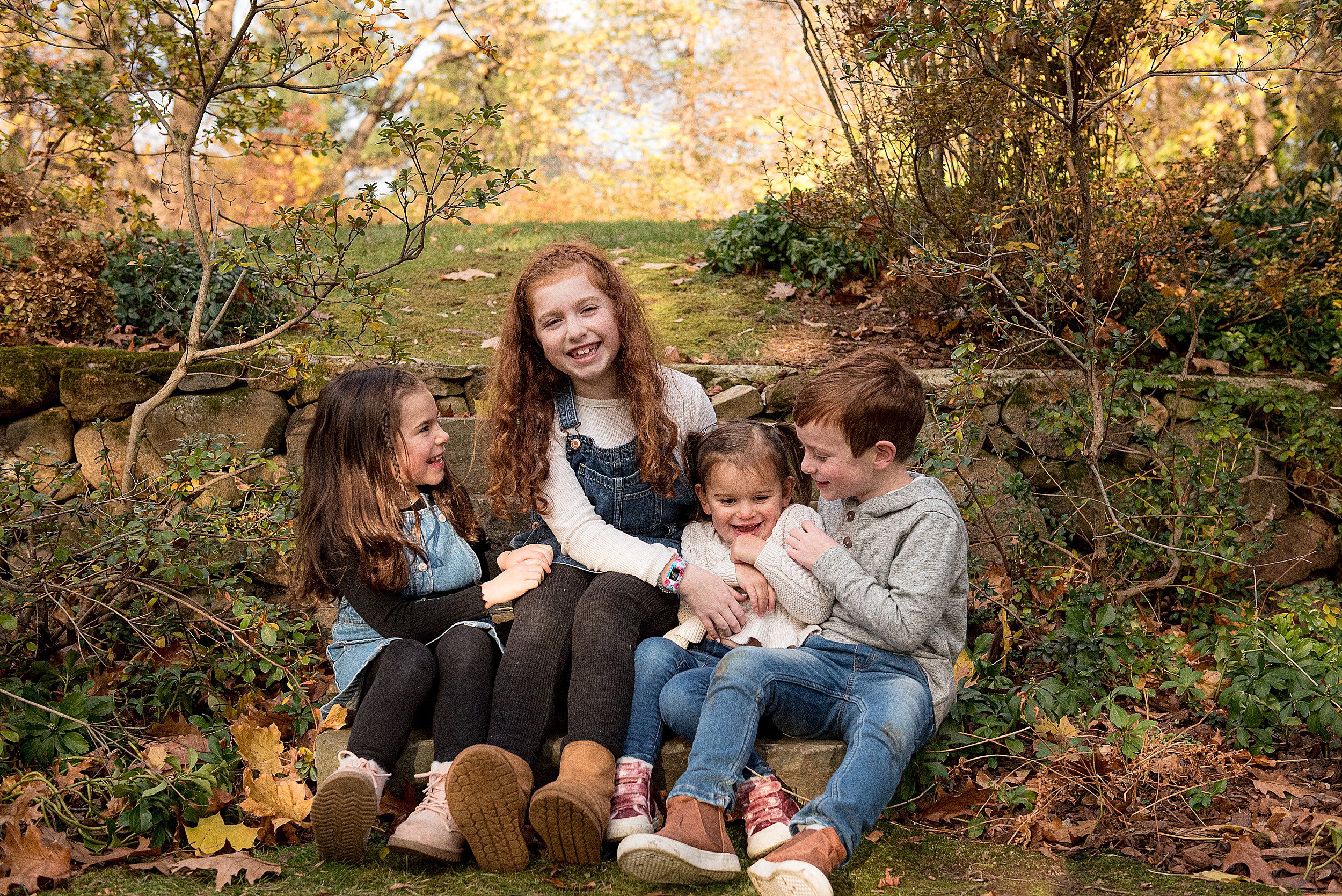 Four siblings sit on a stone step in a garden tickling each other