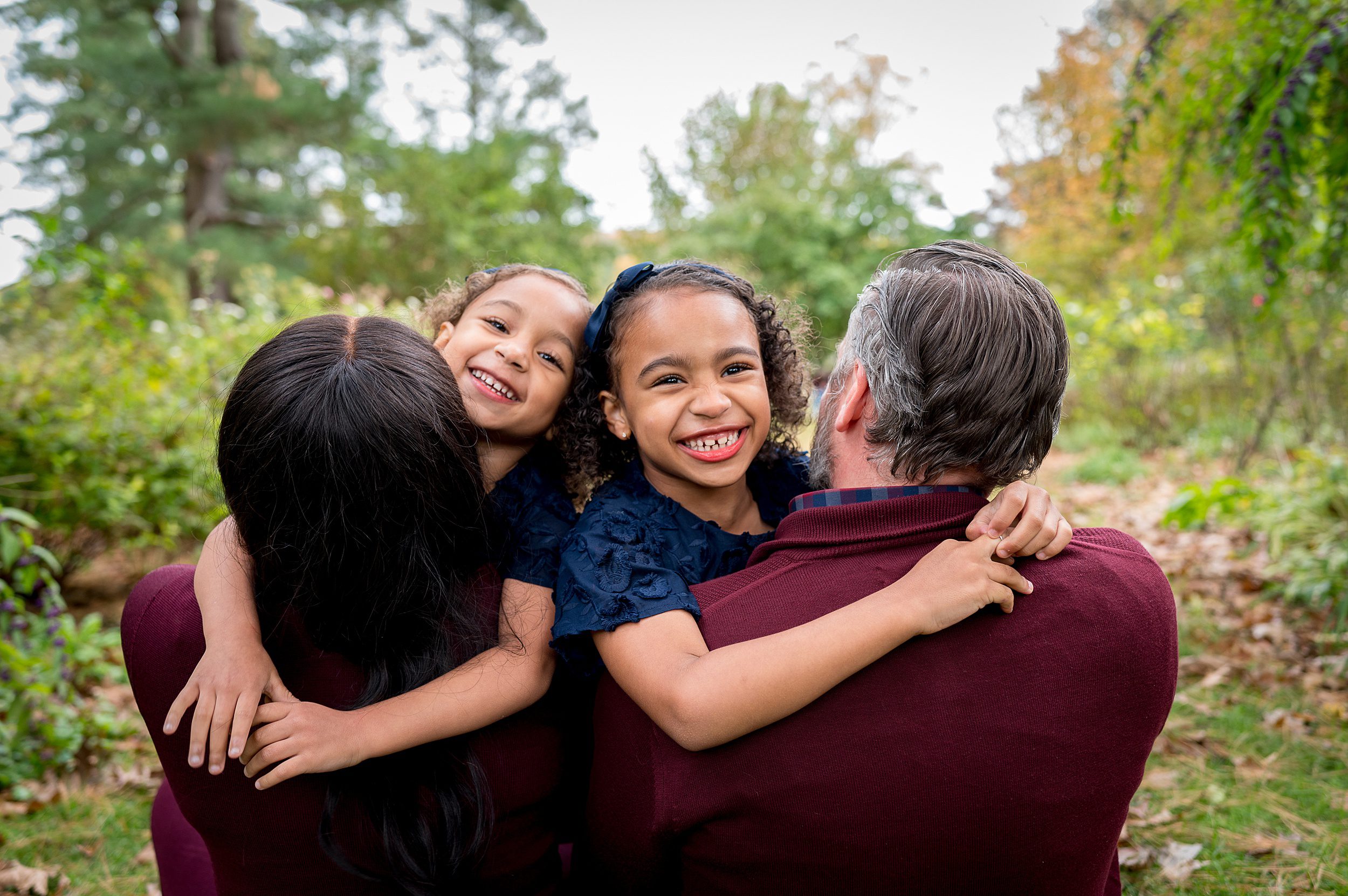 Two young girls climb on and hug their mom and dad in a forest trail wearing matching blue dresses while mom and dad wear maroon sweaters new haven photographer