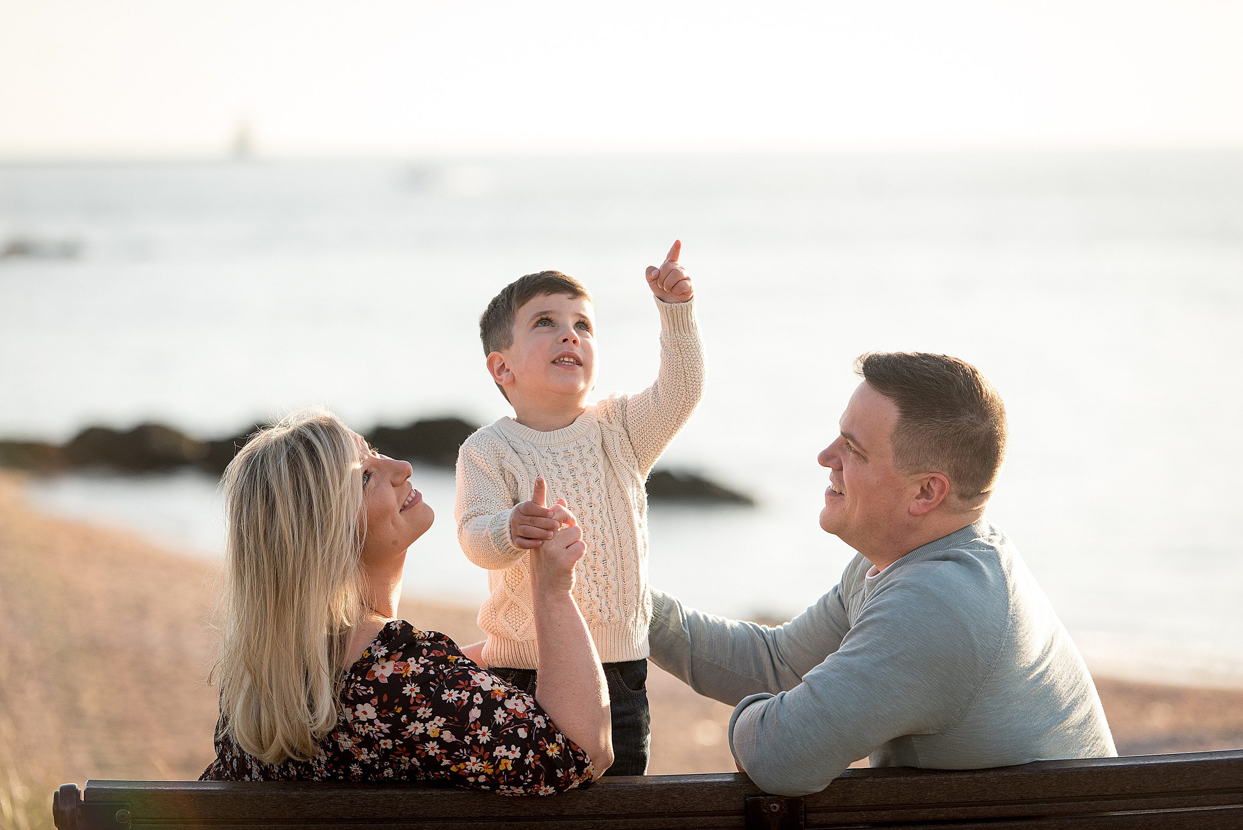 Mom and dad sit on a bench at the beach while playing with their toddler son in a white sweater new haven photographer