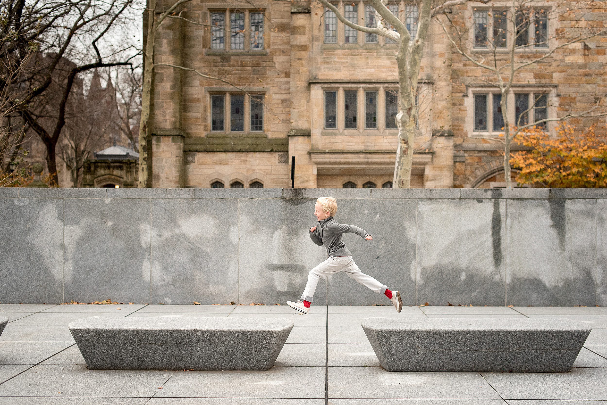 A young boy in white pants and a grey sweat jumps across marble benches in a park new haven photographer