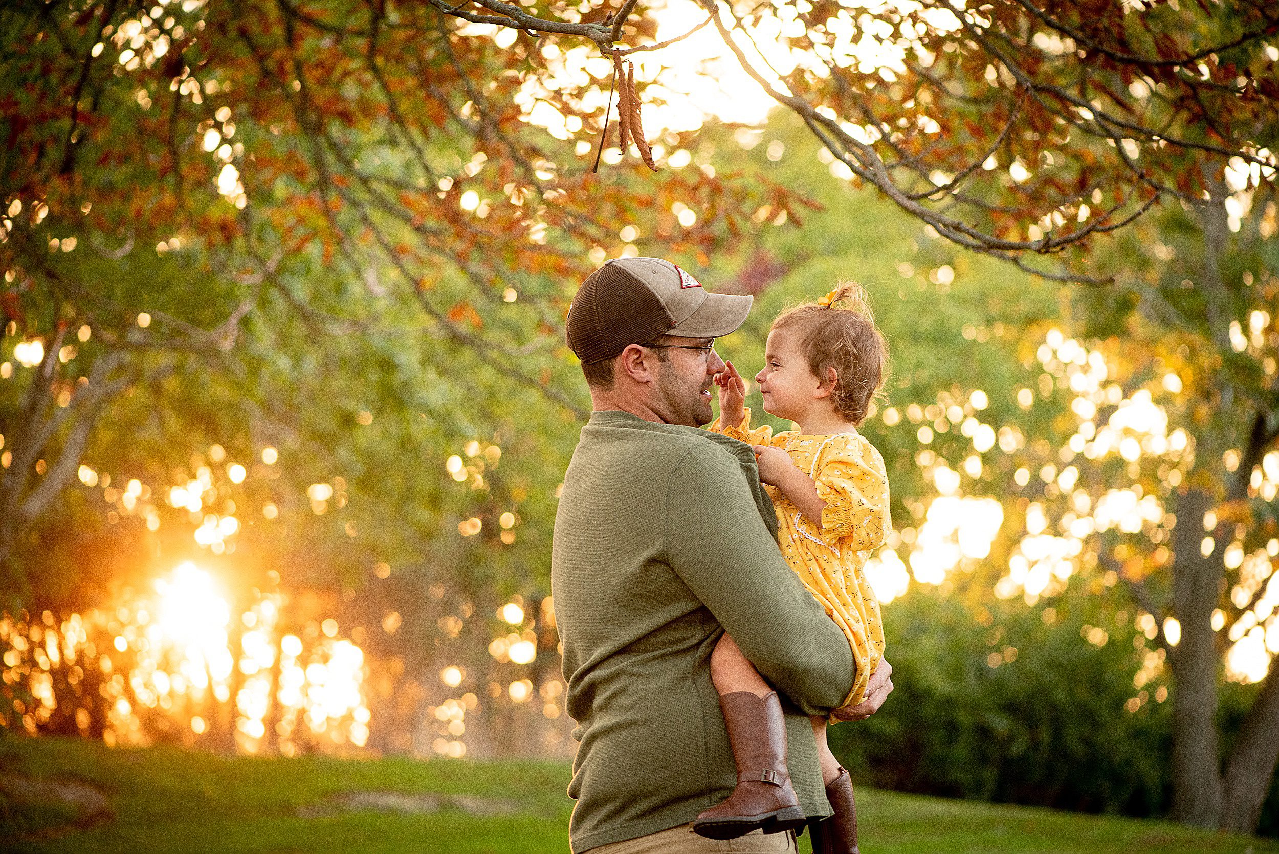 A father in a green shirt stands in a park in fall playing with his toddler daughter in a yellow dress at sunset new haven photographer