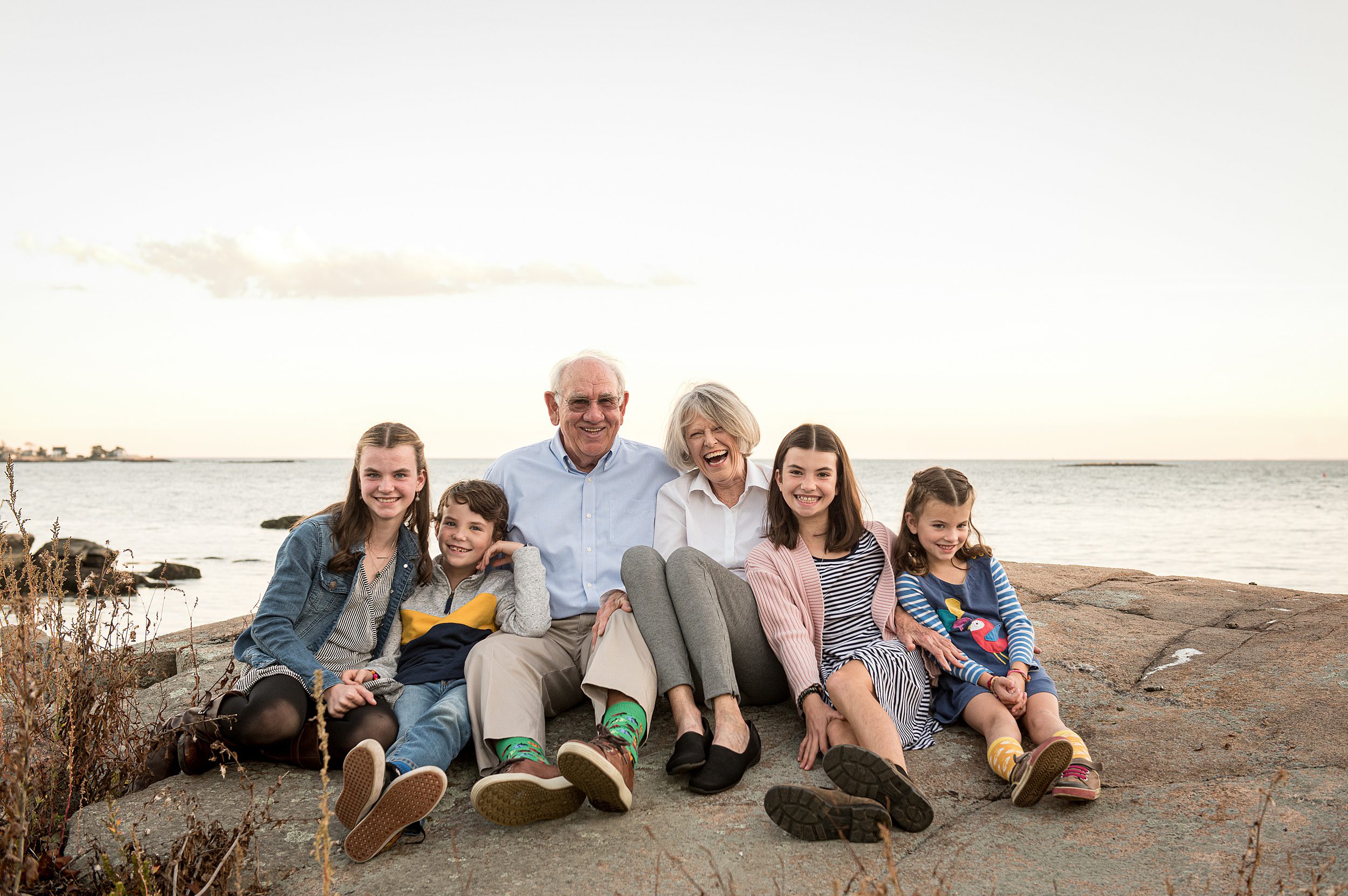 Grandma and grandpa sit on a large rock with their four grand children sitting and cuddling around them on the beach