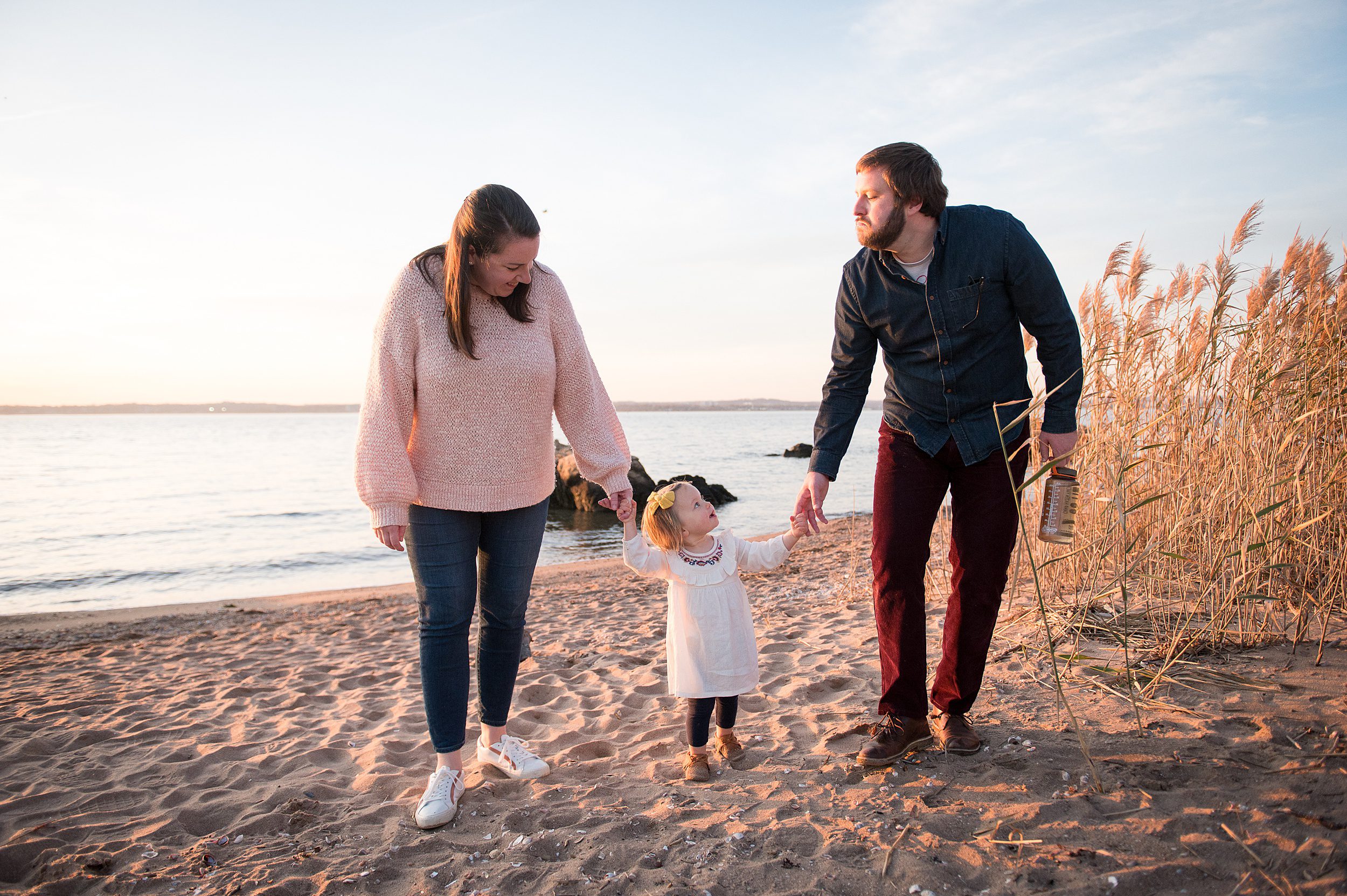 A mom and dad hold hands with their toddler daughter while walking down the best connecticut beaches for families