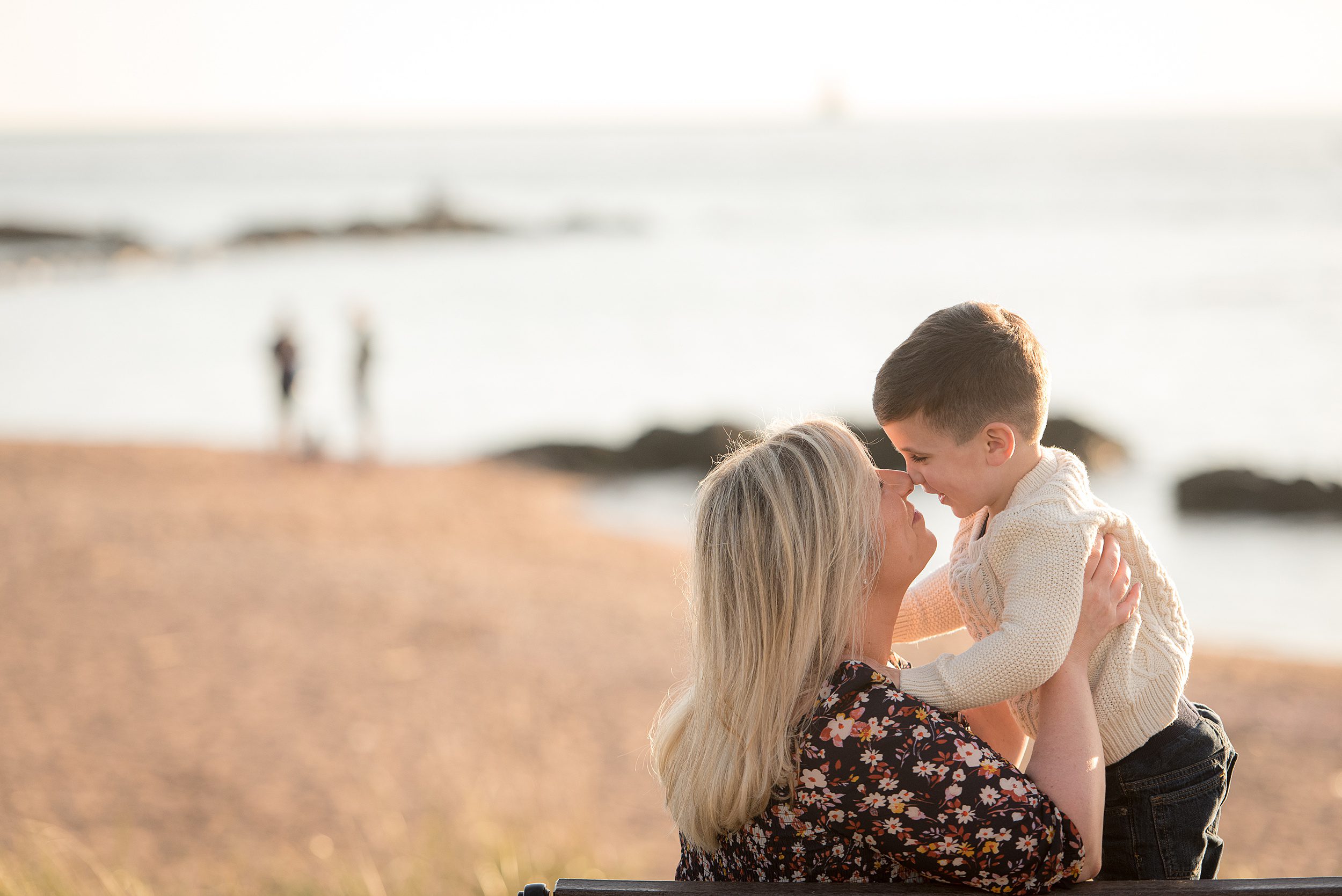 A mother in a black floral dress sits on a bench nuzzling noses with her toddler son in a white sweater at one of the best connecticut beaches for families