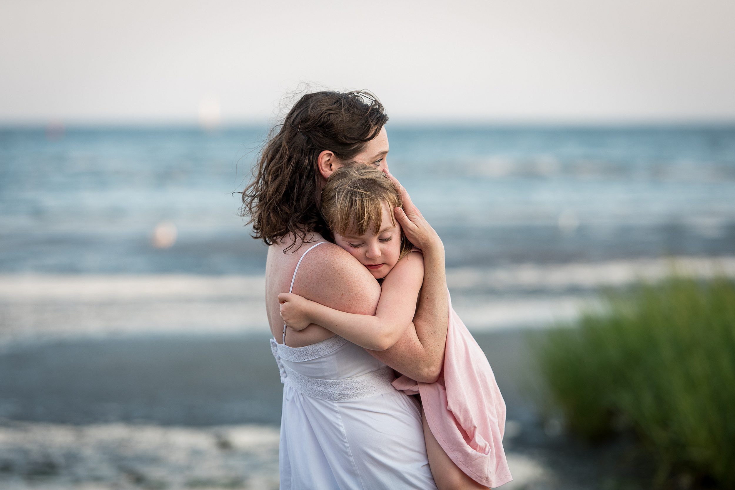 A mother stands on a beach in a white dress cradling her young daughter hugging onto her in a pink dress at one of the best connecticut beaches for families