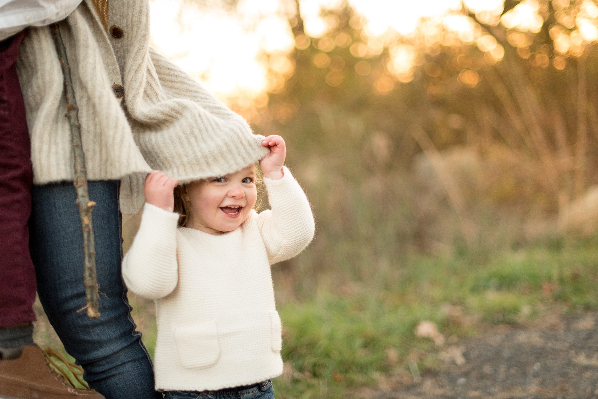 A young girl in a white sweater hides under mom's beige sweater while standing in a park at sunset