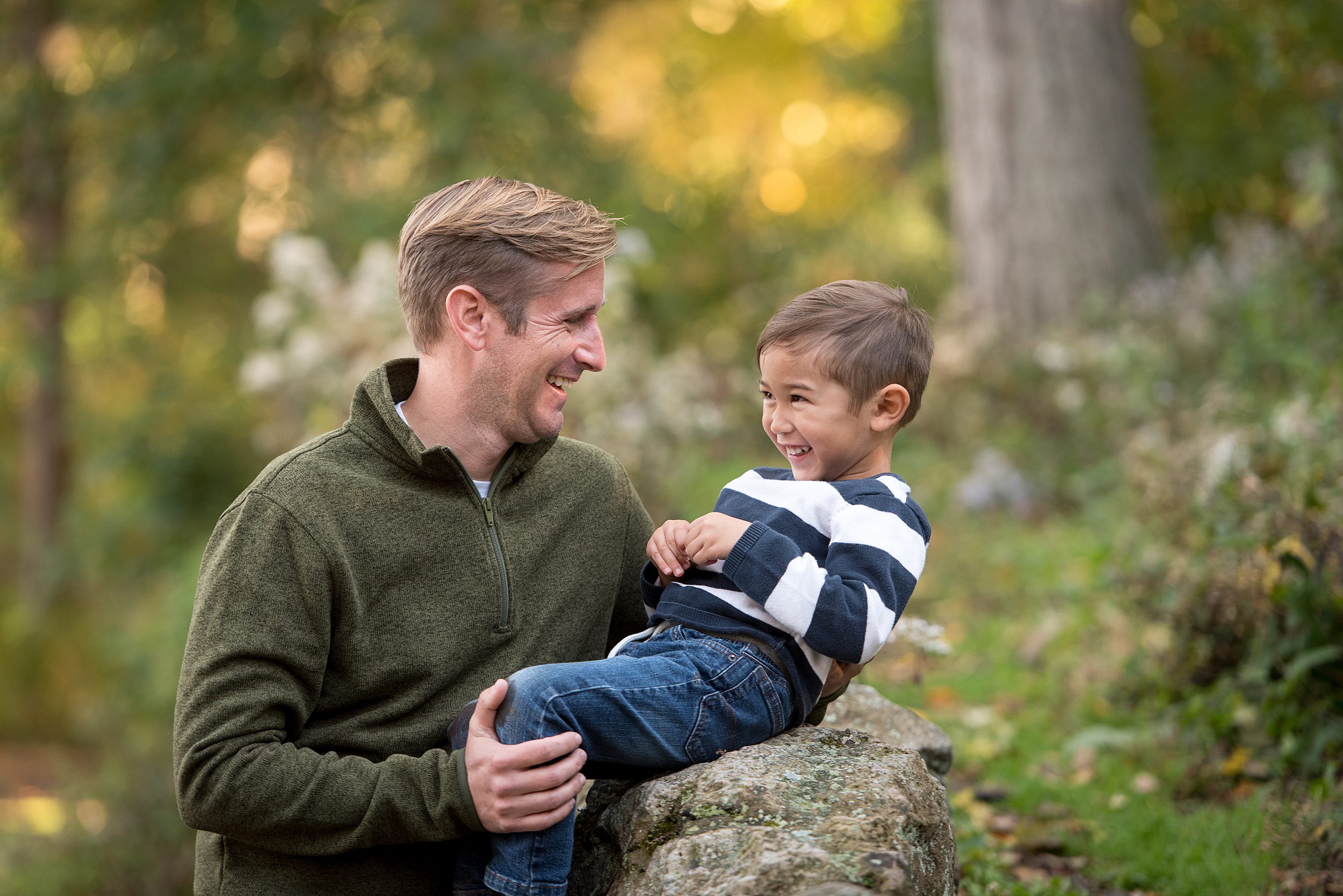 A father plays with his son while he sits on a natural rock wall in a striped shirt