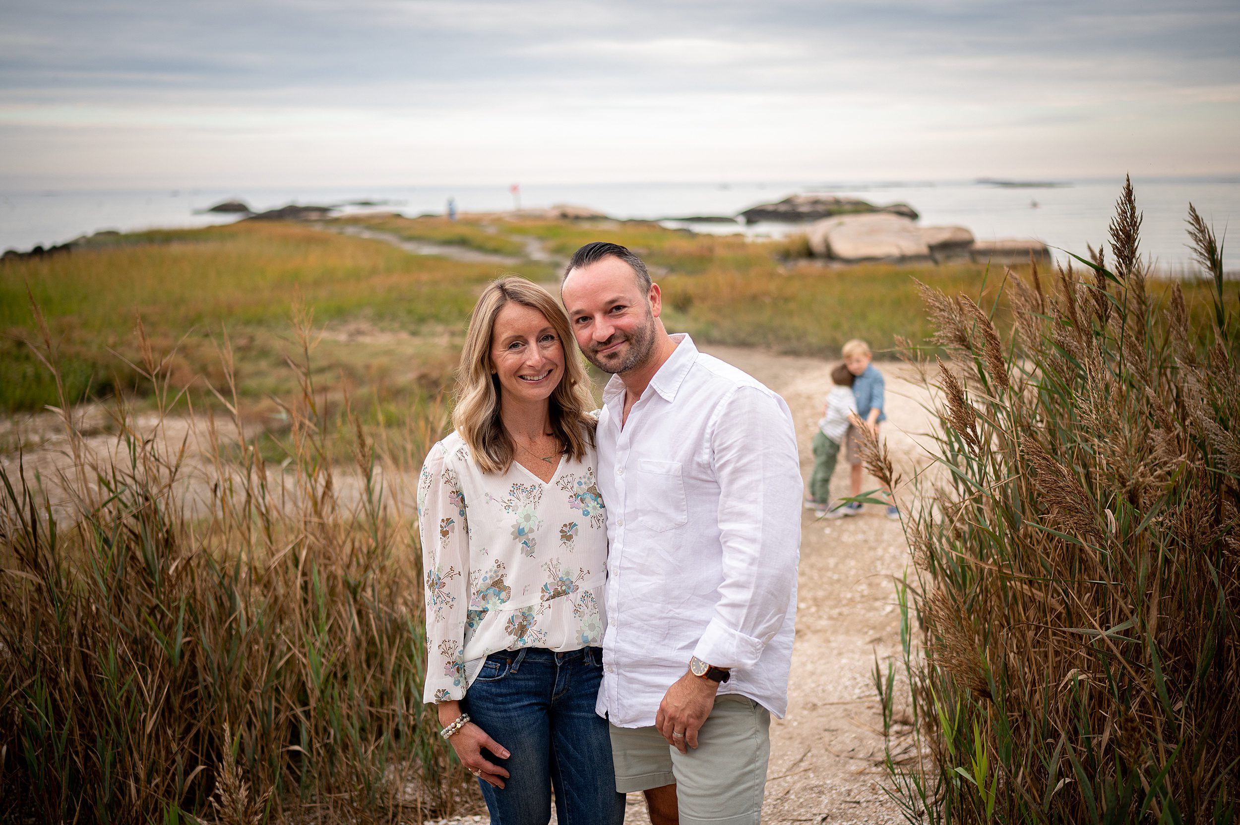 A mother and father smile together while standing in a beach dune path as their two young sons play behind them on the beach after going to a preschool in Madison CT