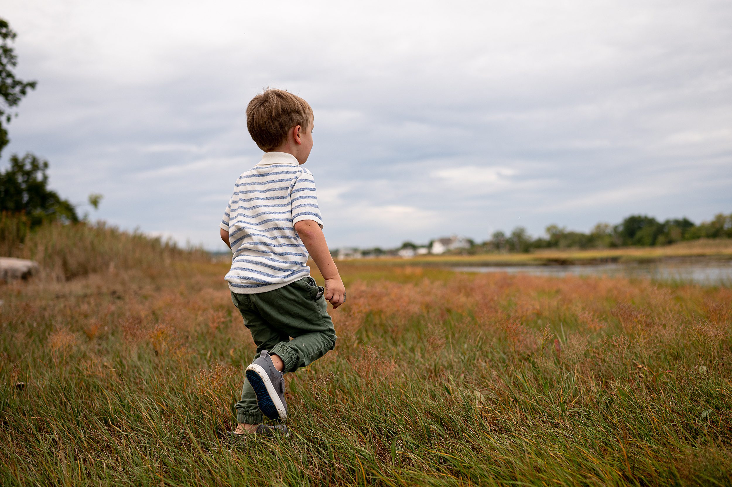A young boy in a striped shirt and green pants runs through a grassy field on the way to a beach