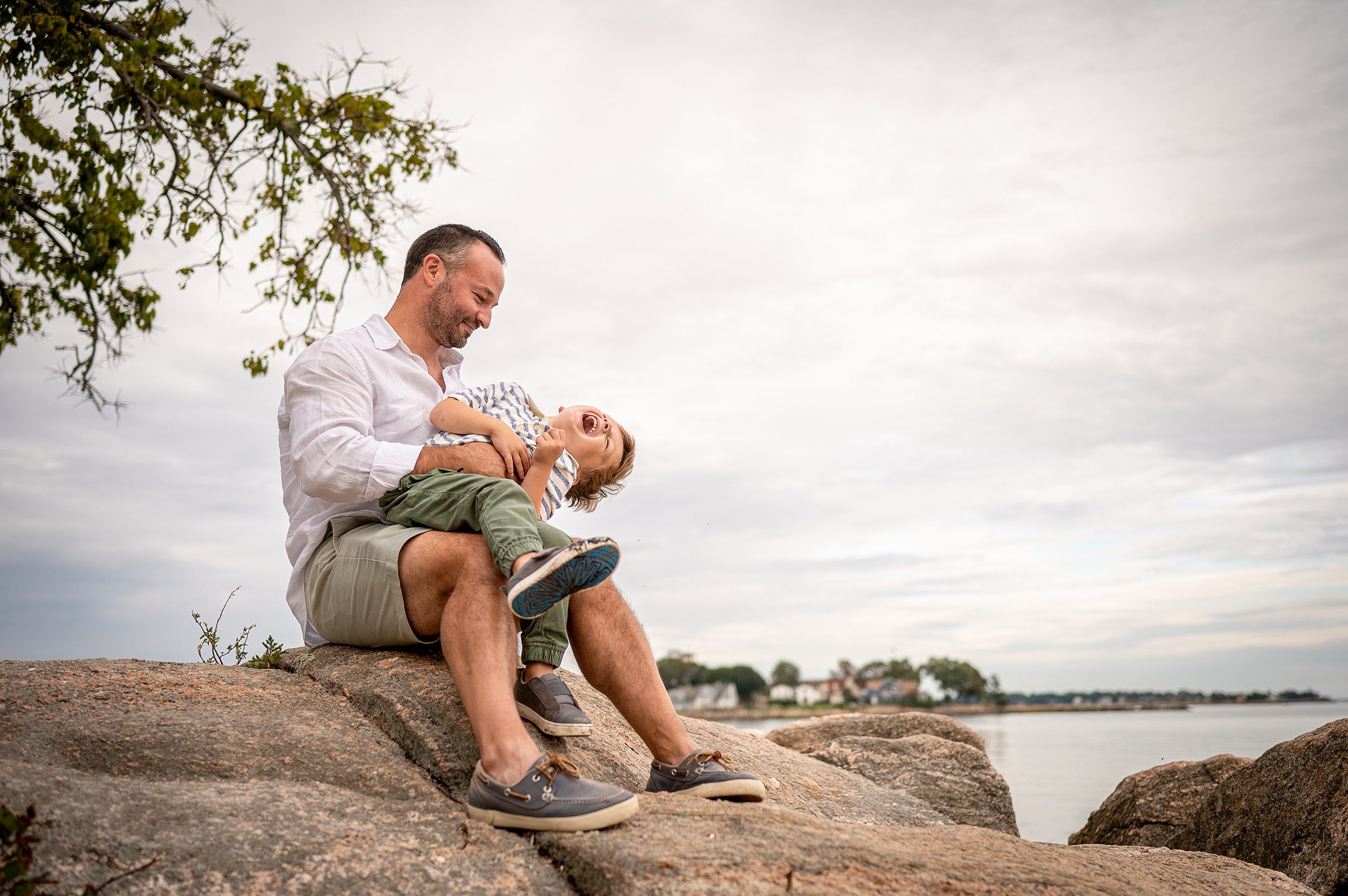 A father sits on a rock in a white shirt on the beach tickling his young son in a green stripe shirt and green pants