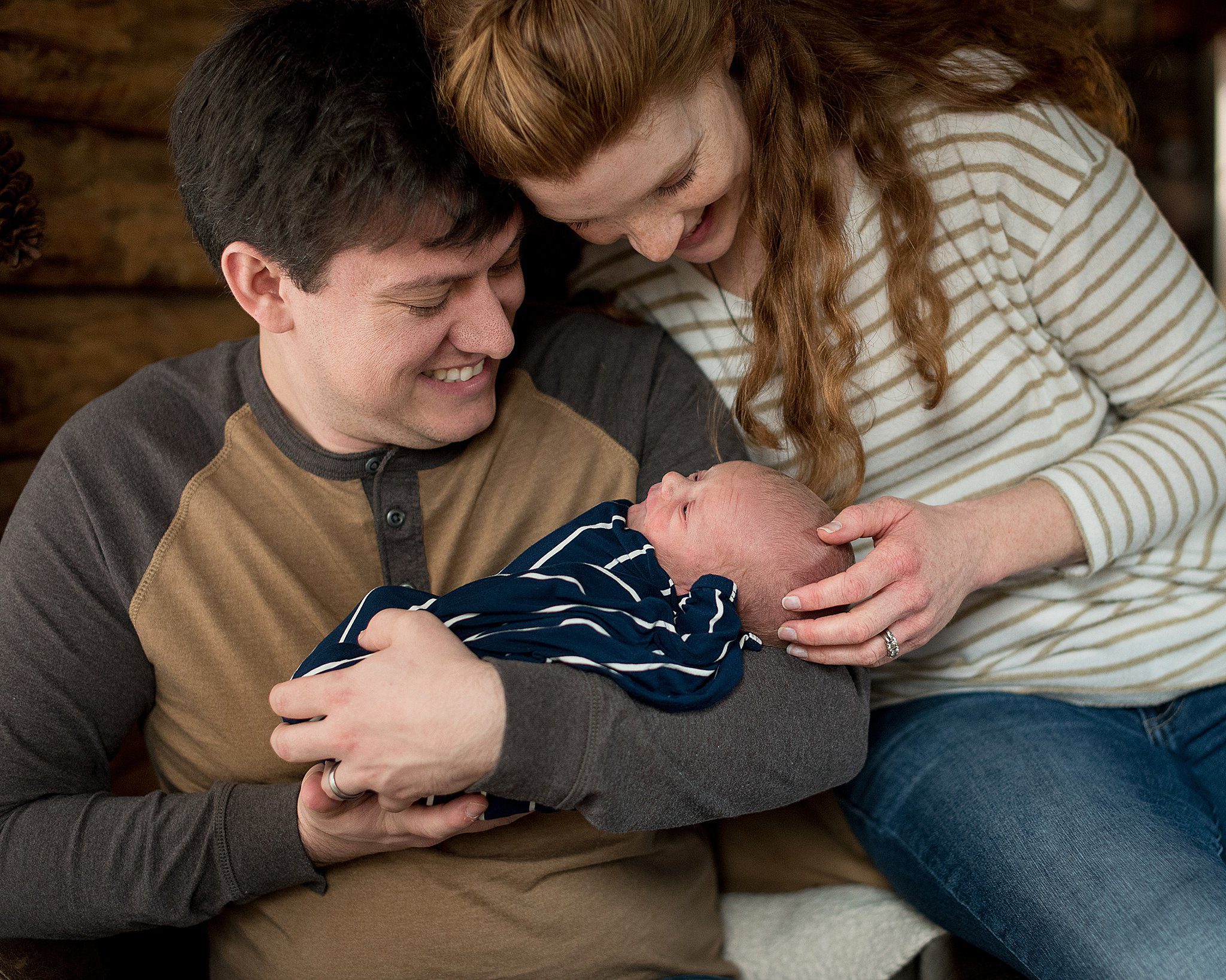 A mom in a stripe shirt leans over her husband cradling their sleeping newborn baby in his arms