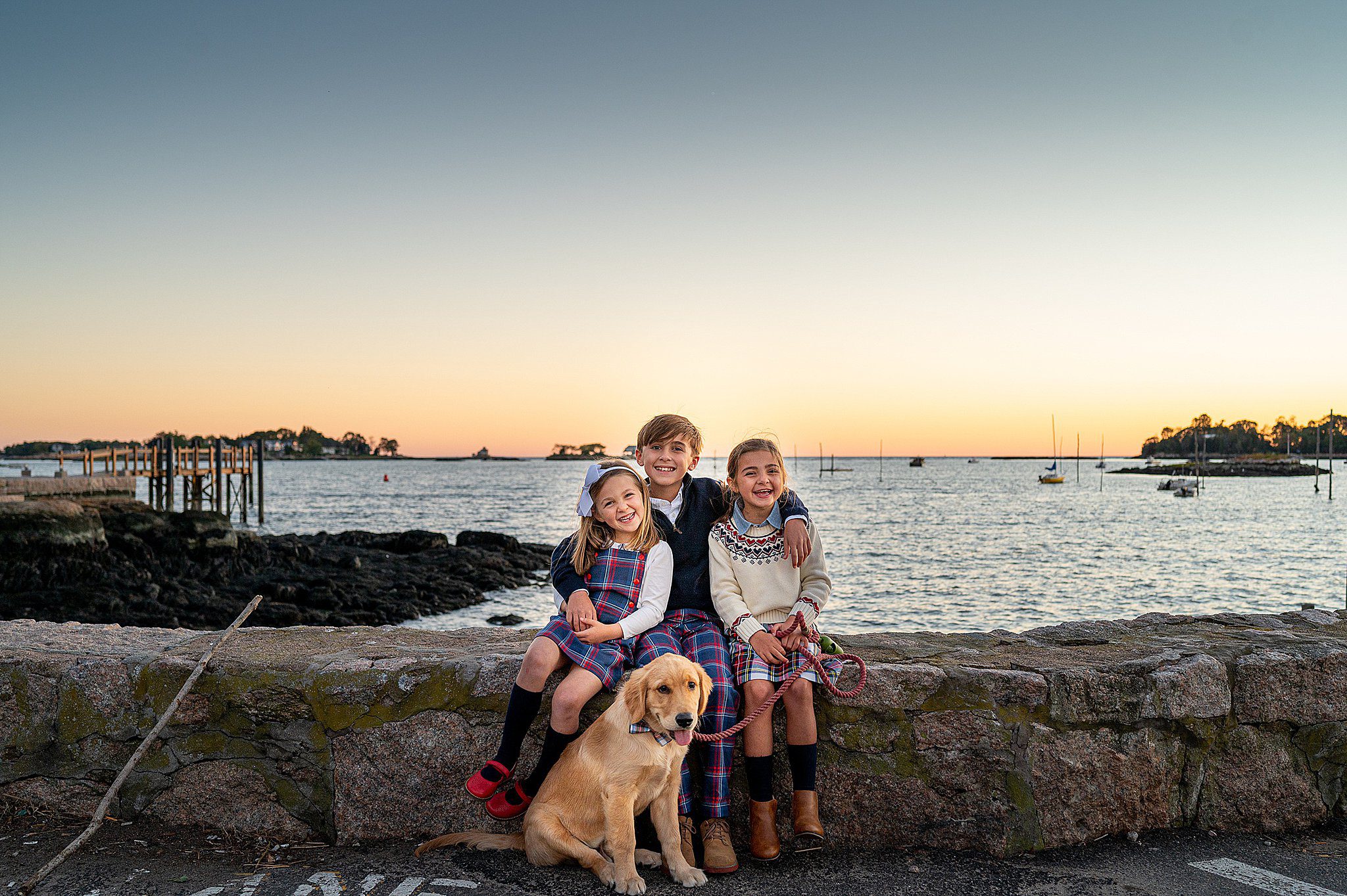 Three siblings sit on a rock wall laughing with their sitting golden retriever in front of them after meeting babysitters connecticut