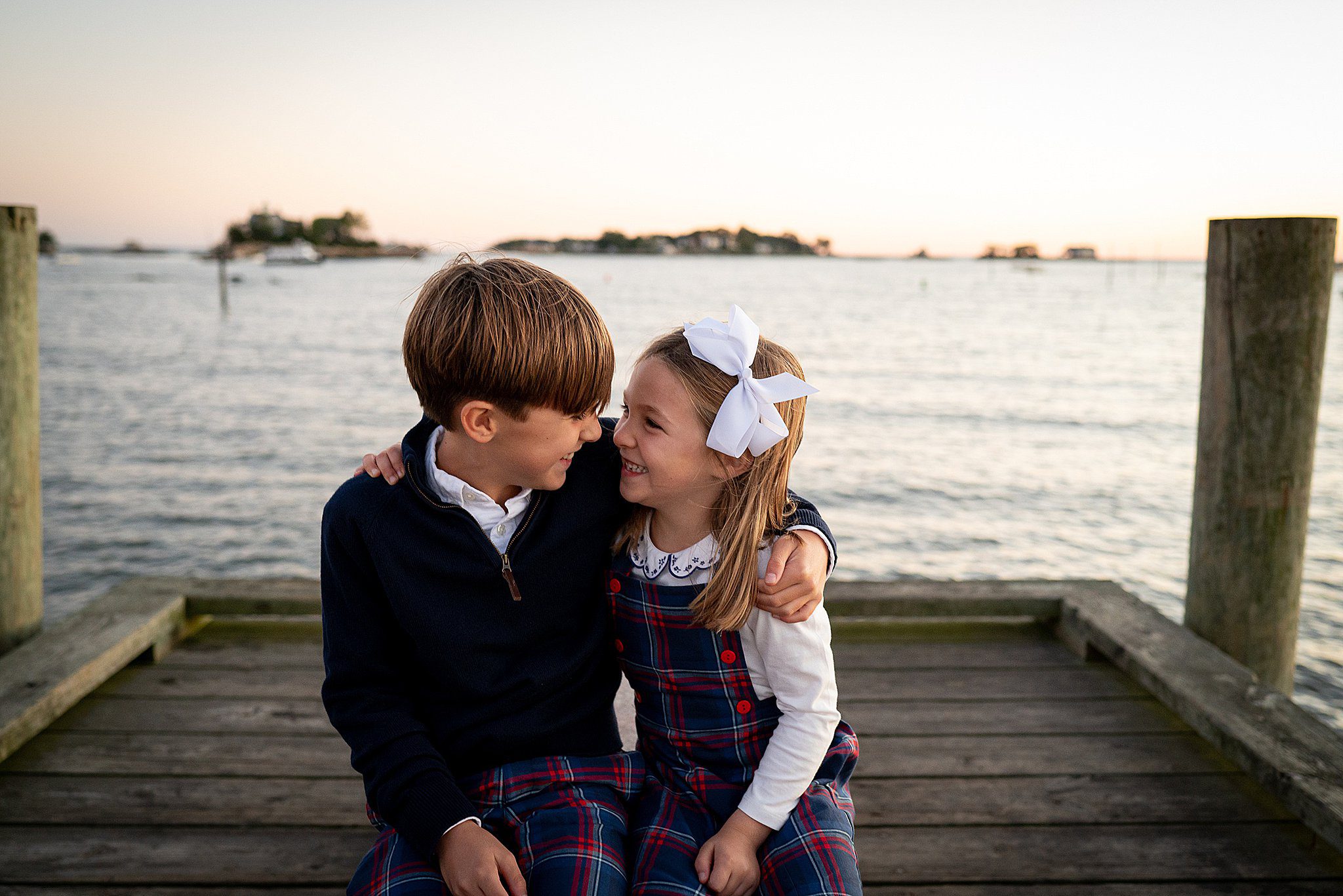 Young siblings giggle while holding each other close and sitting on a dock in matching plaid outfits after meeting babysitters connecticut