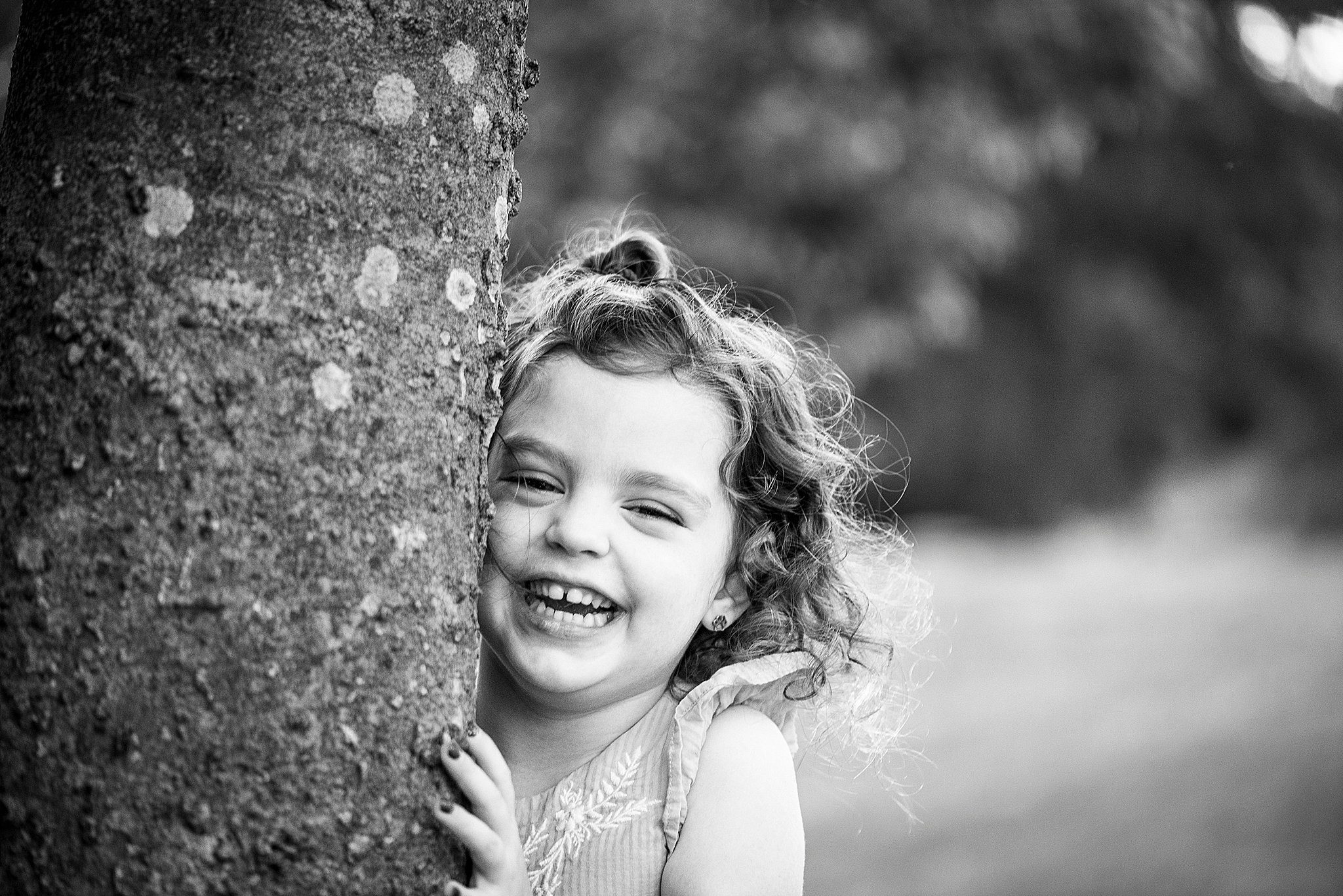 A toddler girl laughs while hiding behind a large tree
