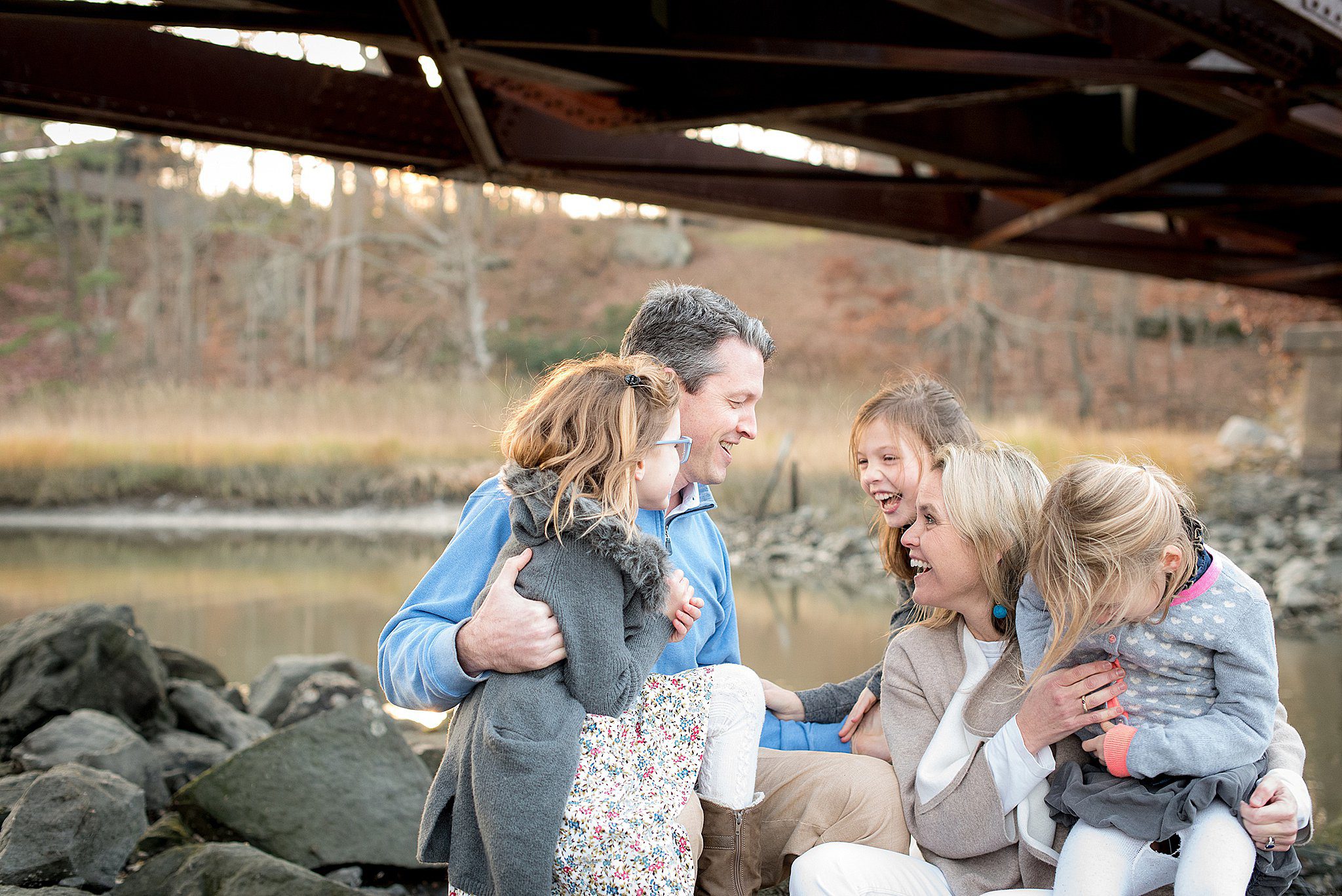 A family of five with three young daughter laughs and plays on rocks along a river under a bridge