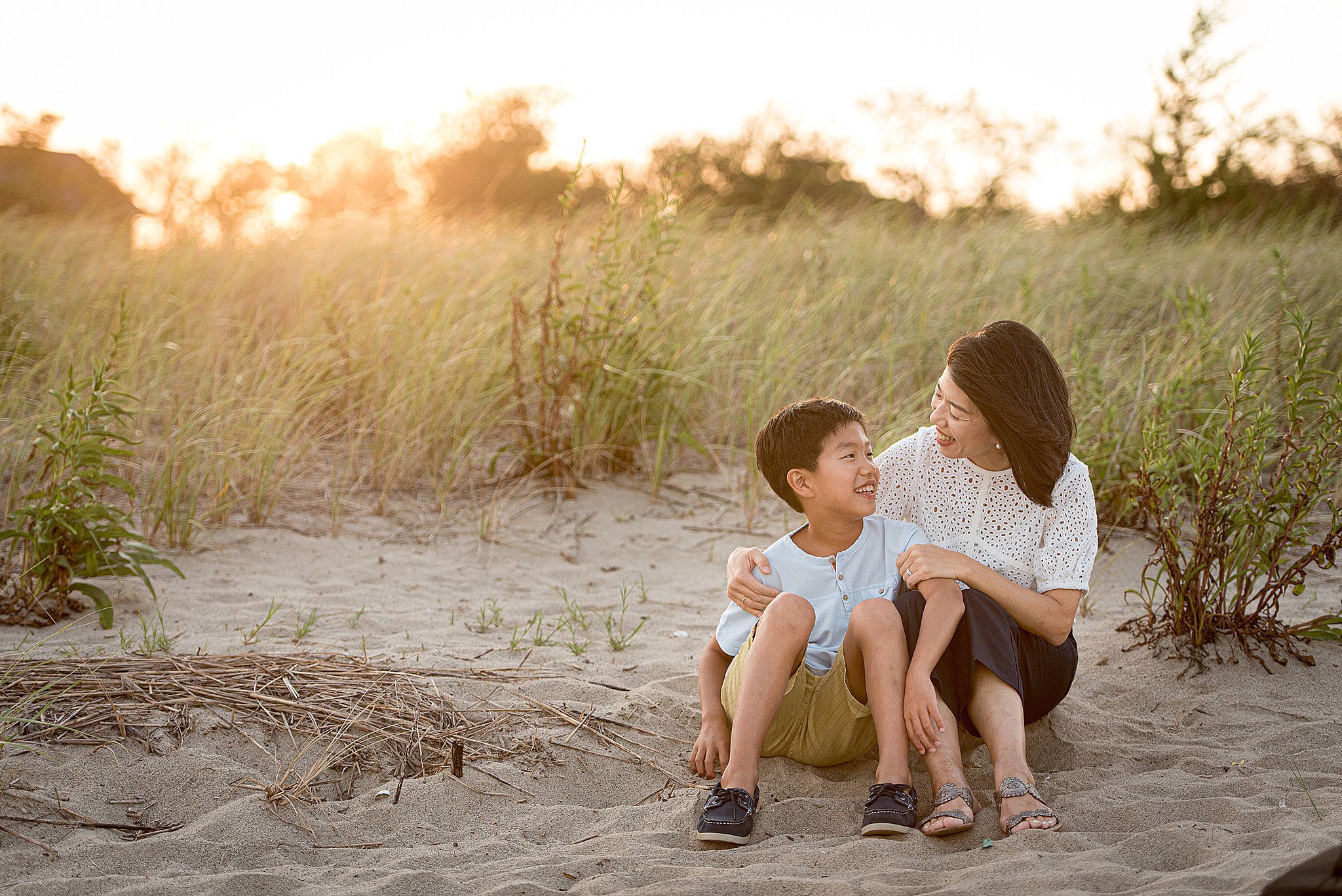 A mother in a white top sits in the sand on a beach with her young son at sunset