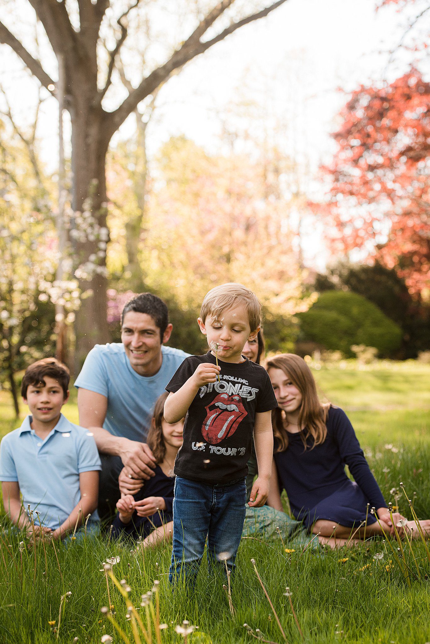 A young boy in a shirt and jeans blows dandelions while his family sits in the grass behind him