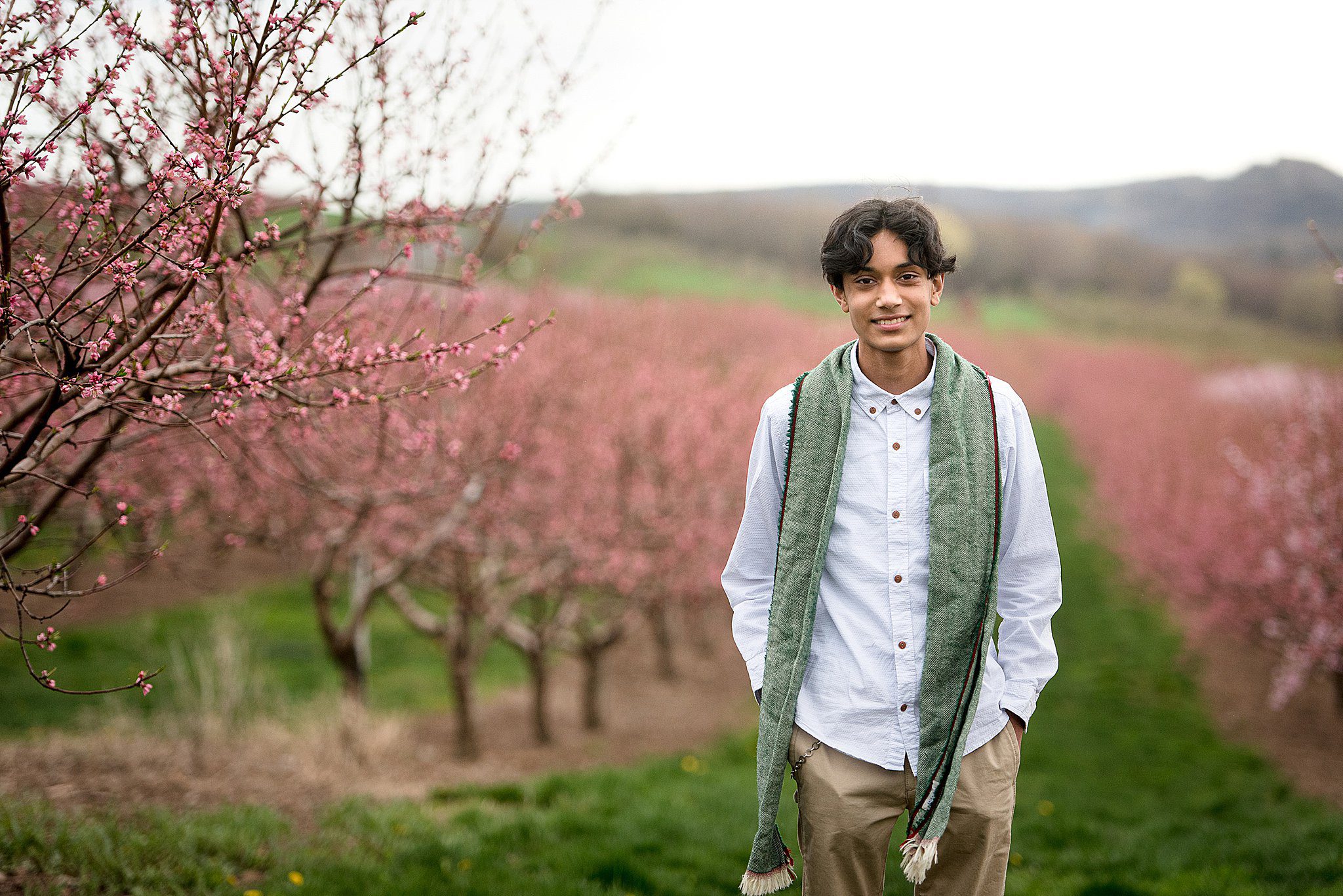 A man in a white shirt and green scarf stands in an orchard of pink blooming trees with hands in pockets during things to do in guilford ct