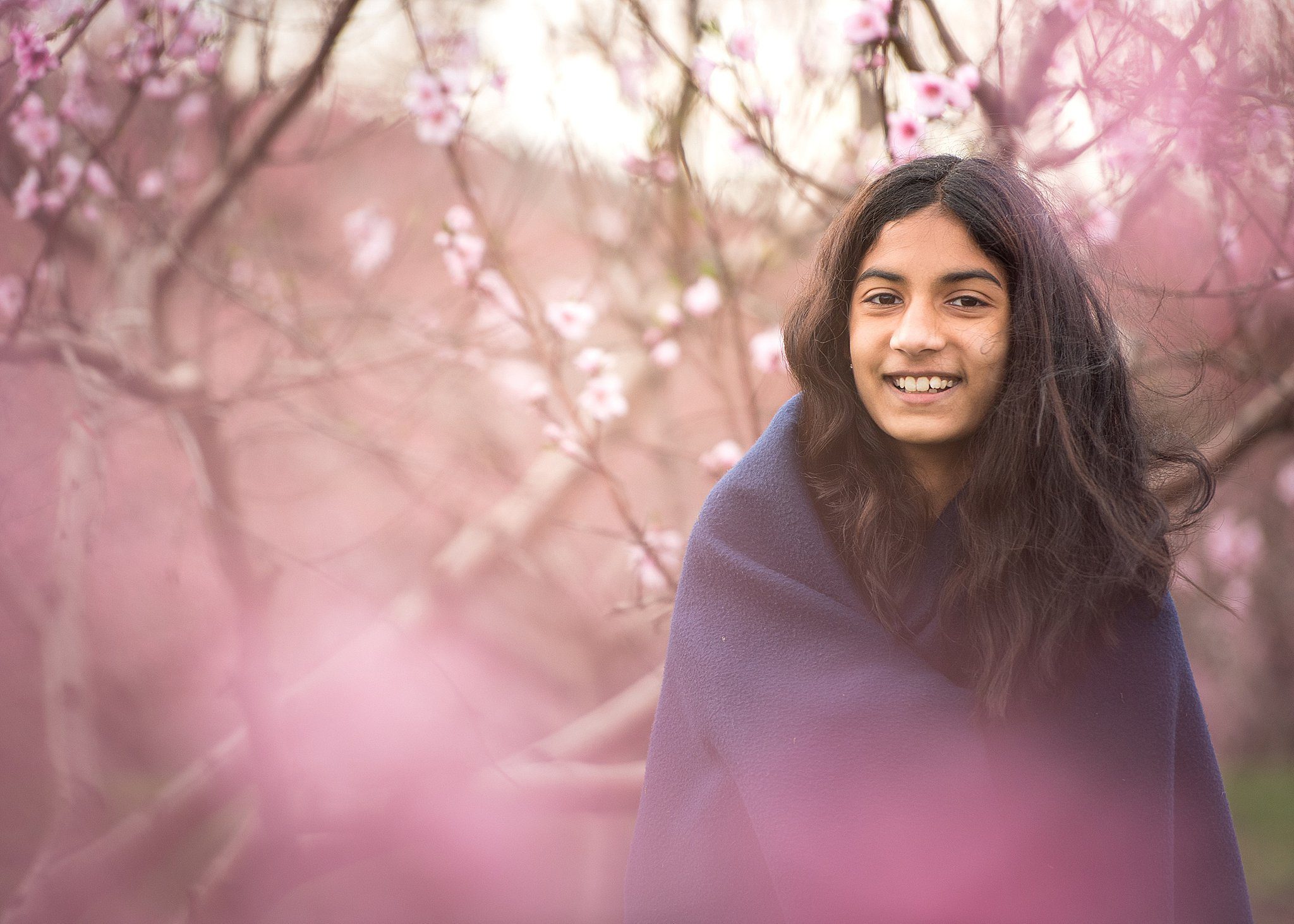 A young girl wrapped in a blue blanket stands amongst pink blooming trees