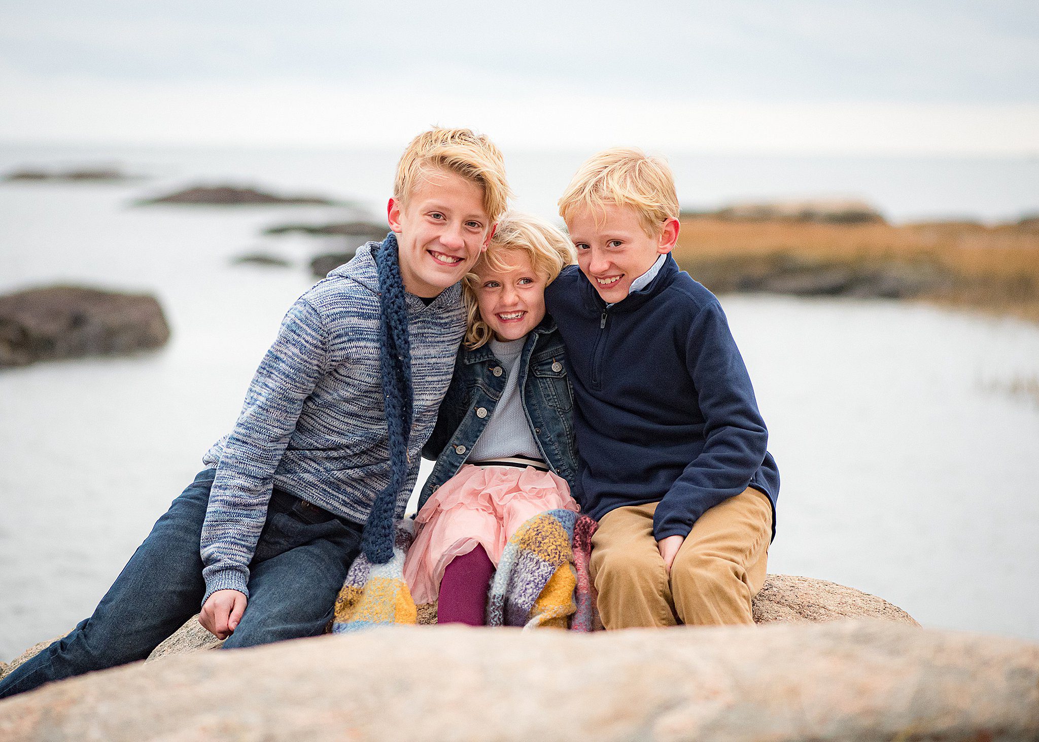 Three blonde and smiling siblings sit on a large rock on the water hugging before visiting toy store guilford ct