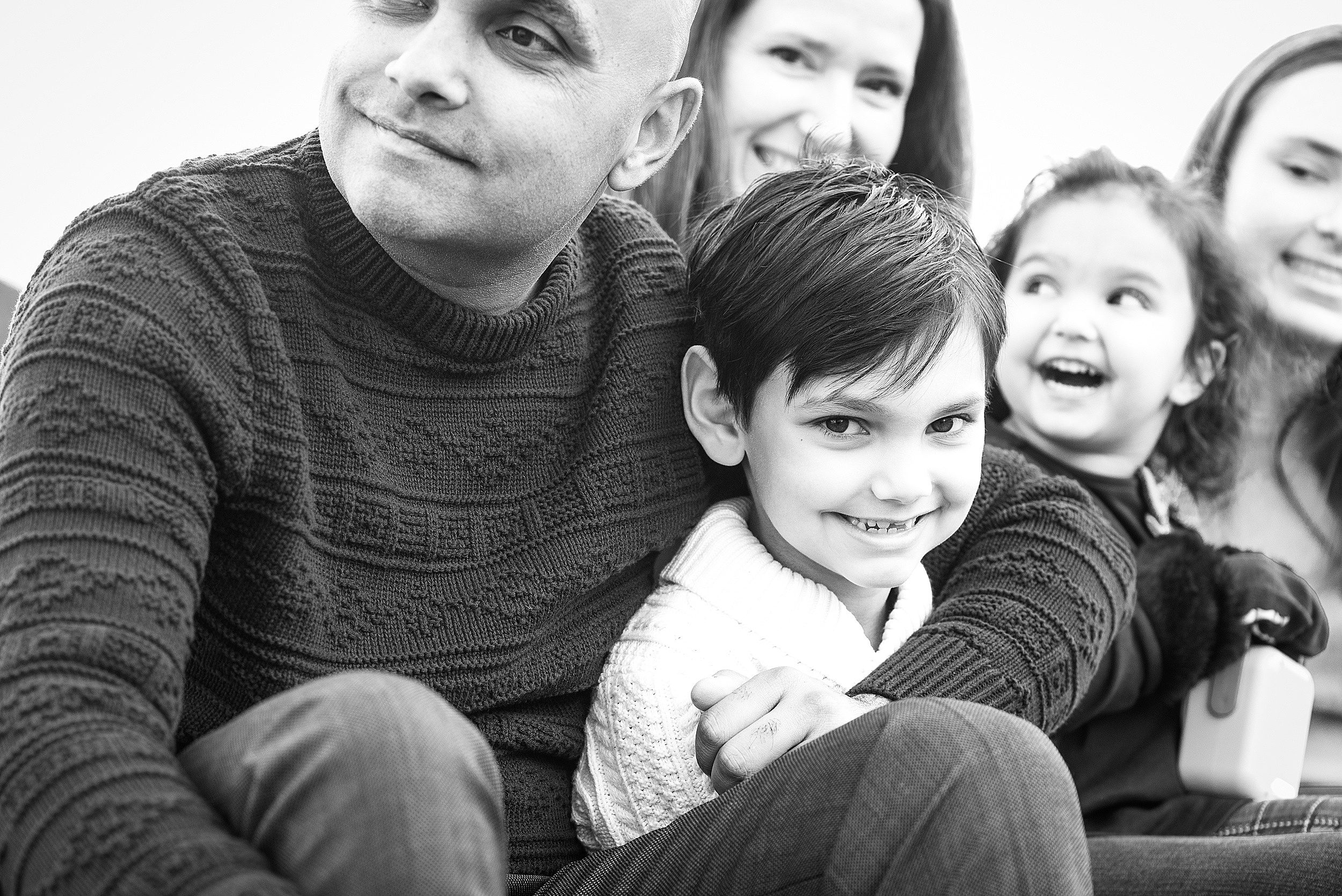 A toddler boy sits in dad's lap being hugged with his family behind them