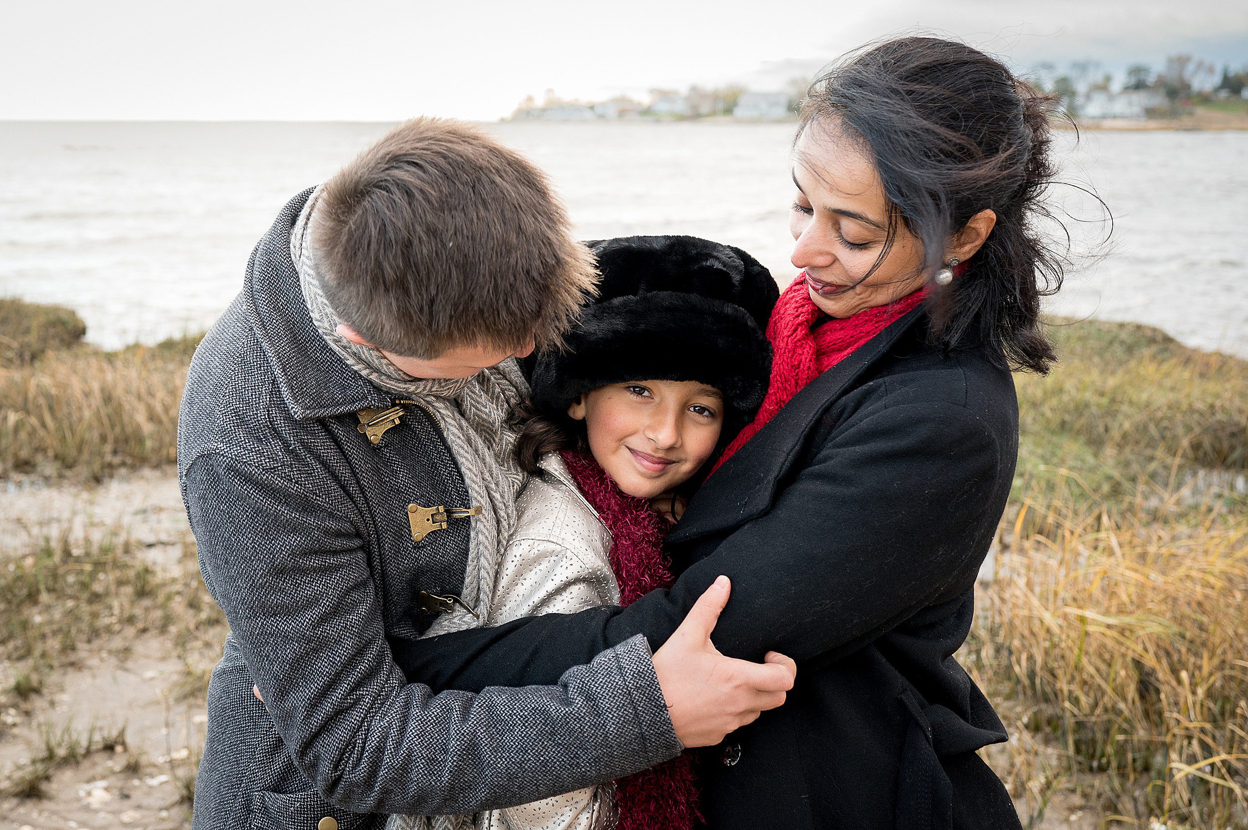 A mom and dad hug their young daughter while standing on a beach in fall after meting pediatrician madison ct