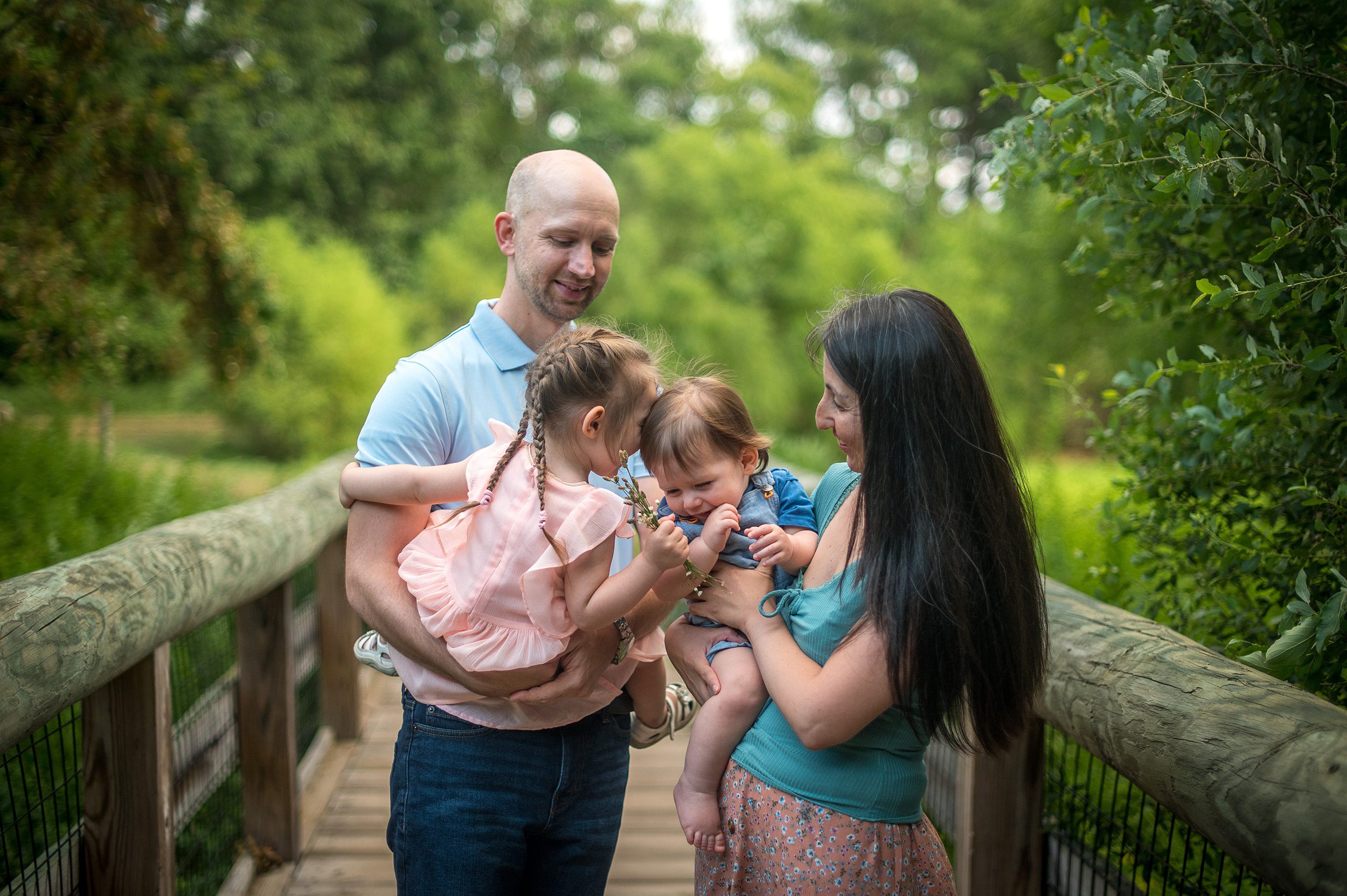 A mom and dad play with their toddler son and daughter on a wooden bridge while picking wildflowers before visiting the connecticut children's museum
