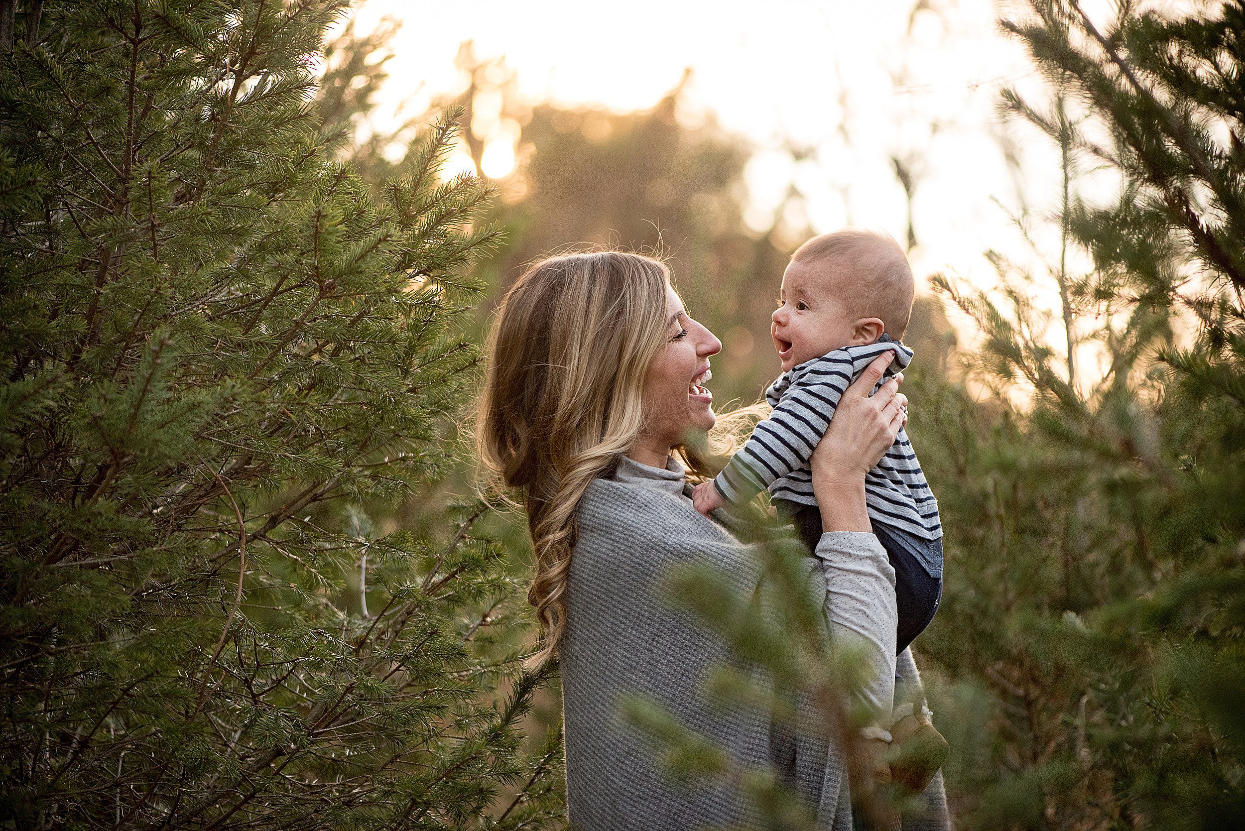 A happy mother in a grey sweater laughs with her toddler son in a pine forest at sunset