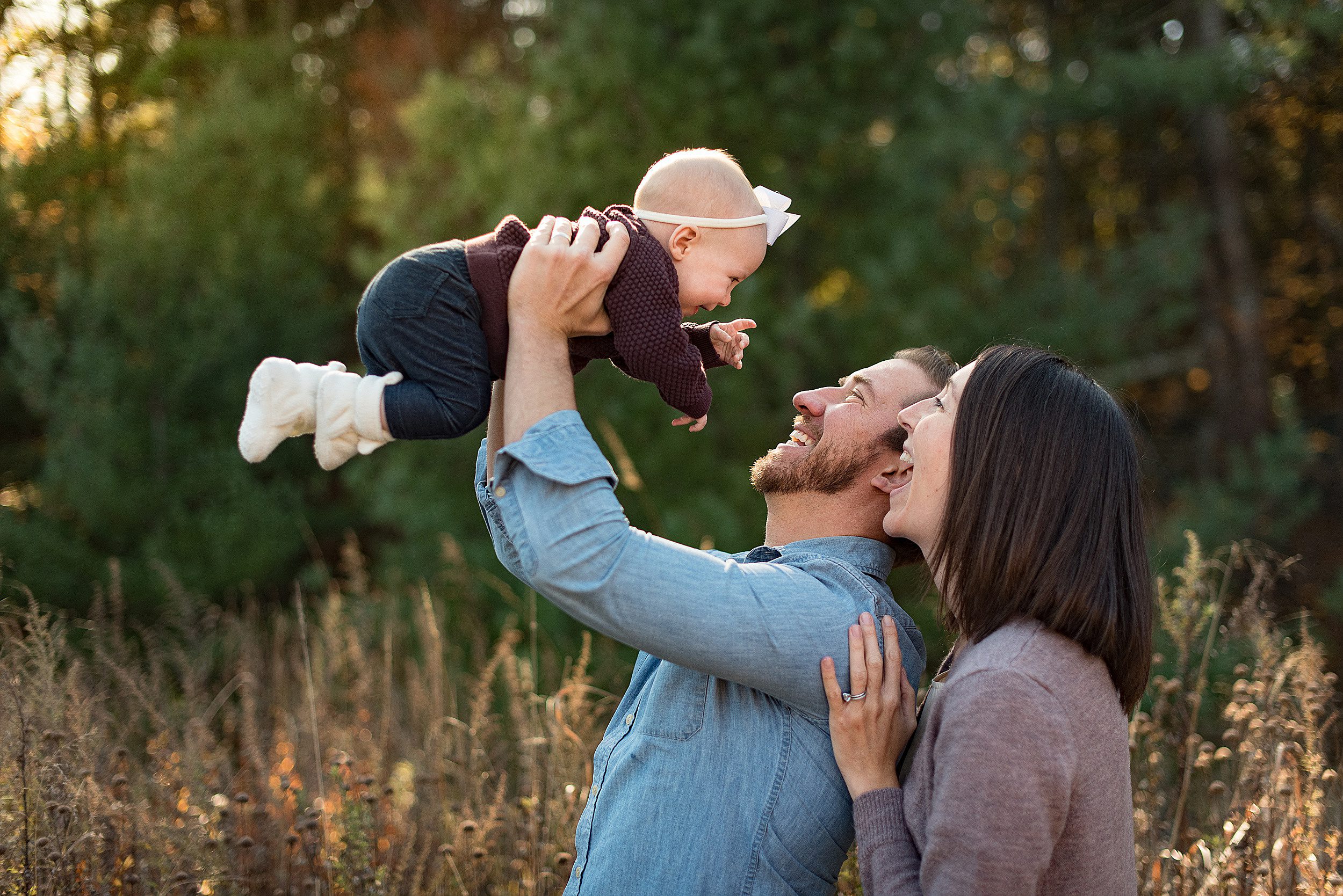 A happy mom and dad lift and play with their toddler daugher in a park at sunset