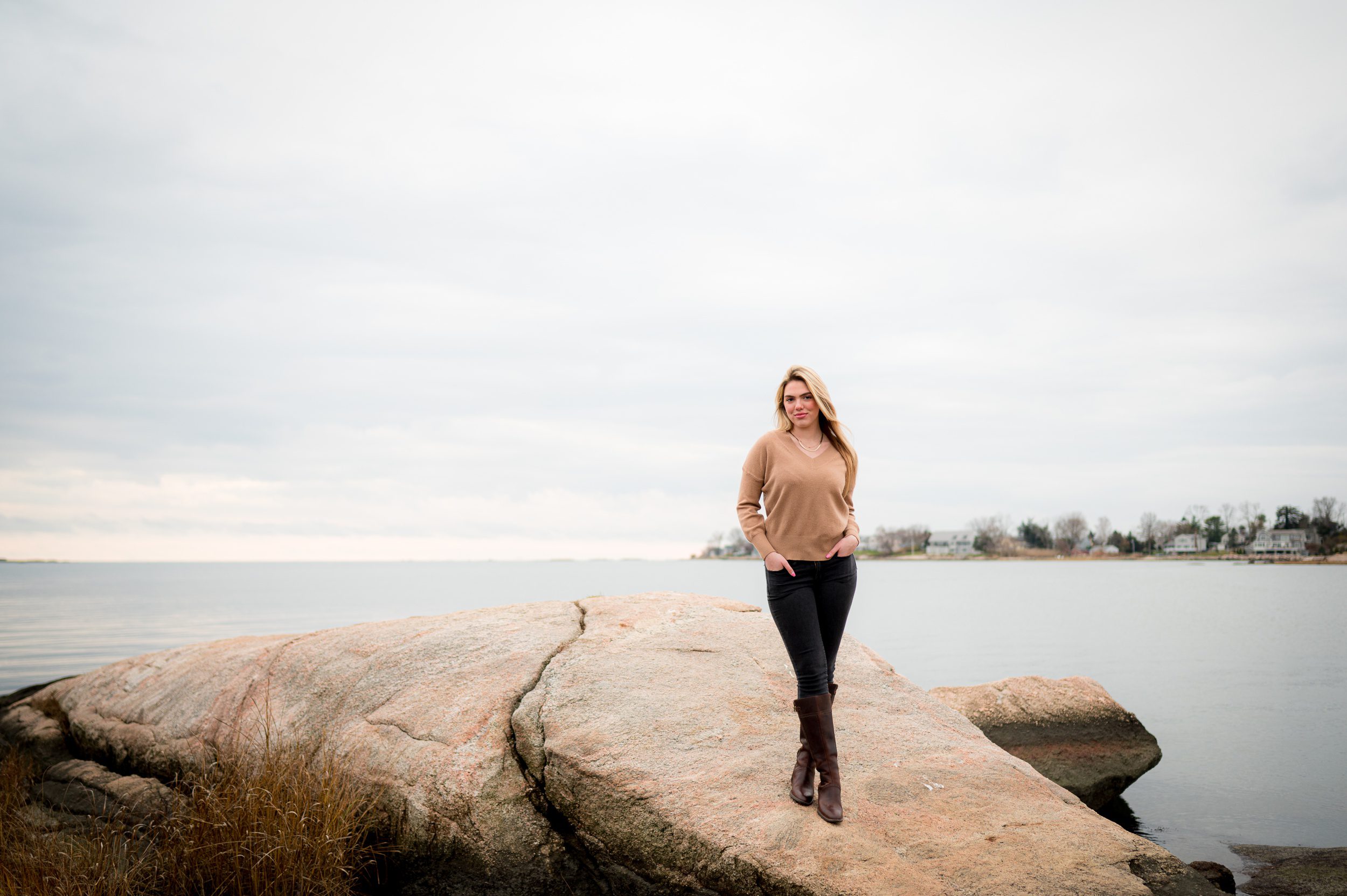 A high school senior in black pants and brown shirt walks on a large boulder on the water