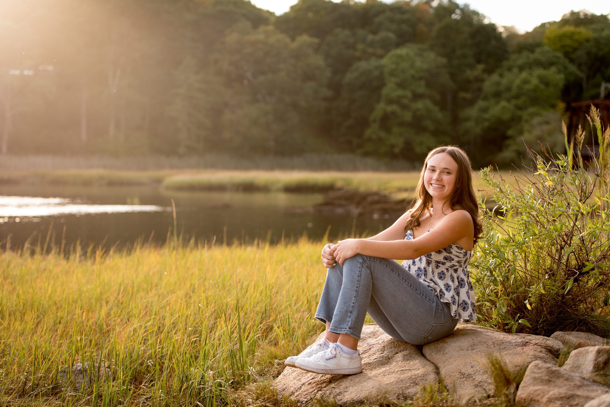 A happy high school senior sits on a rock by the water in jeans and blue blouse after attending a college fair in connecticut