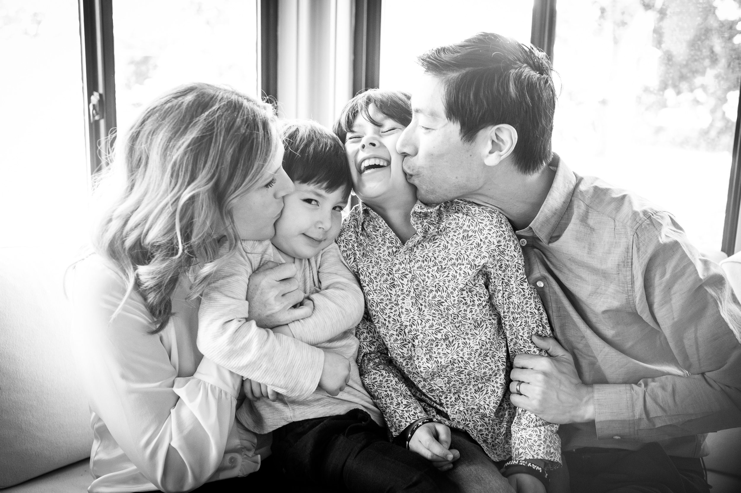 Happy toddlers are kissed by mom and dad on the cheeks while sitting in their laps on a couch after attending guilford preschools