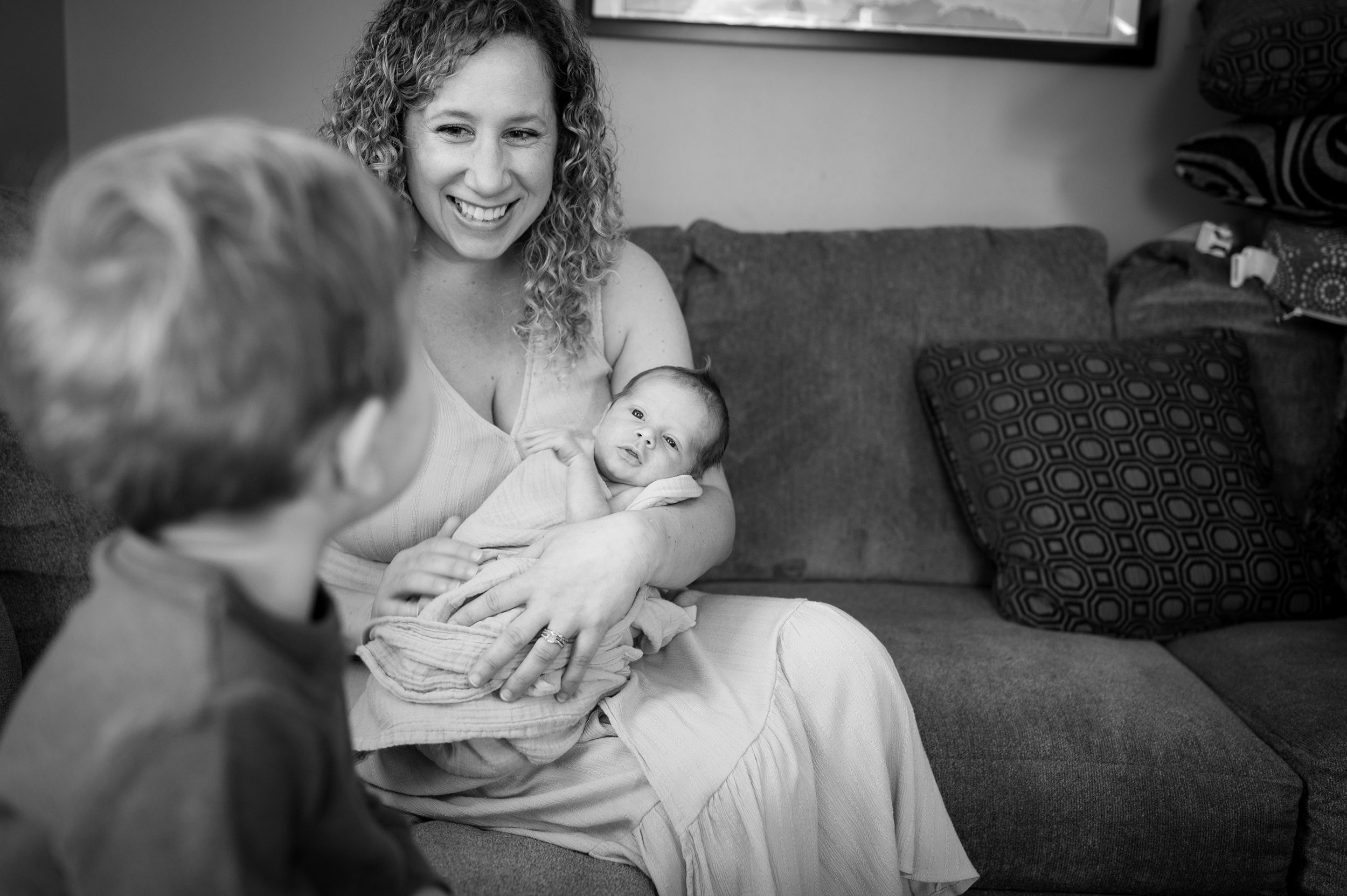 A mom smiles big while holding her newborn on a couch looking at her toddler son