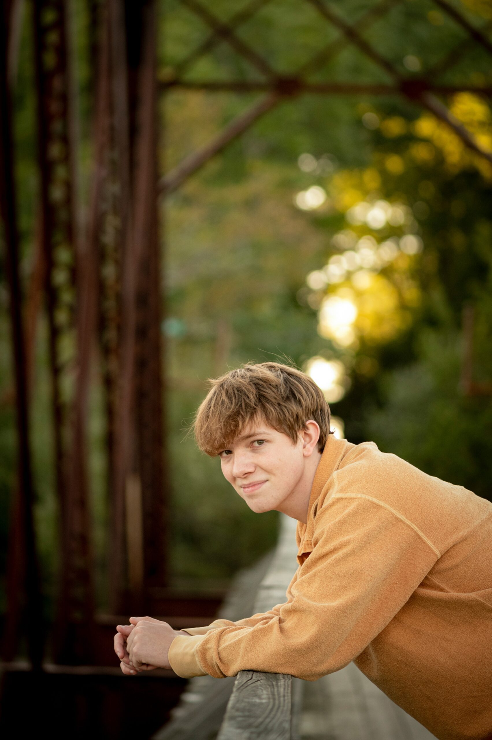 A high school senior leans on a bridge railing at sunset in an orange shirt
