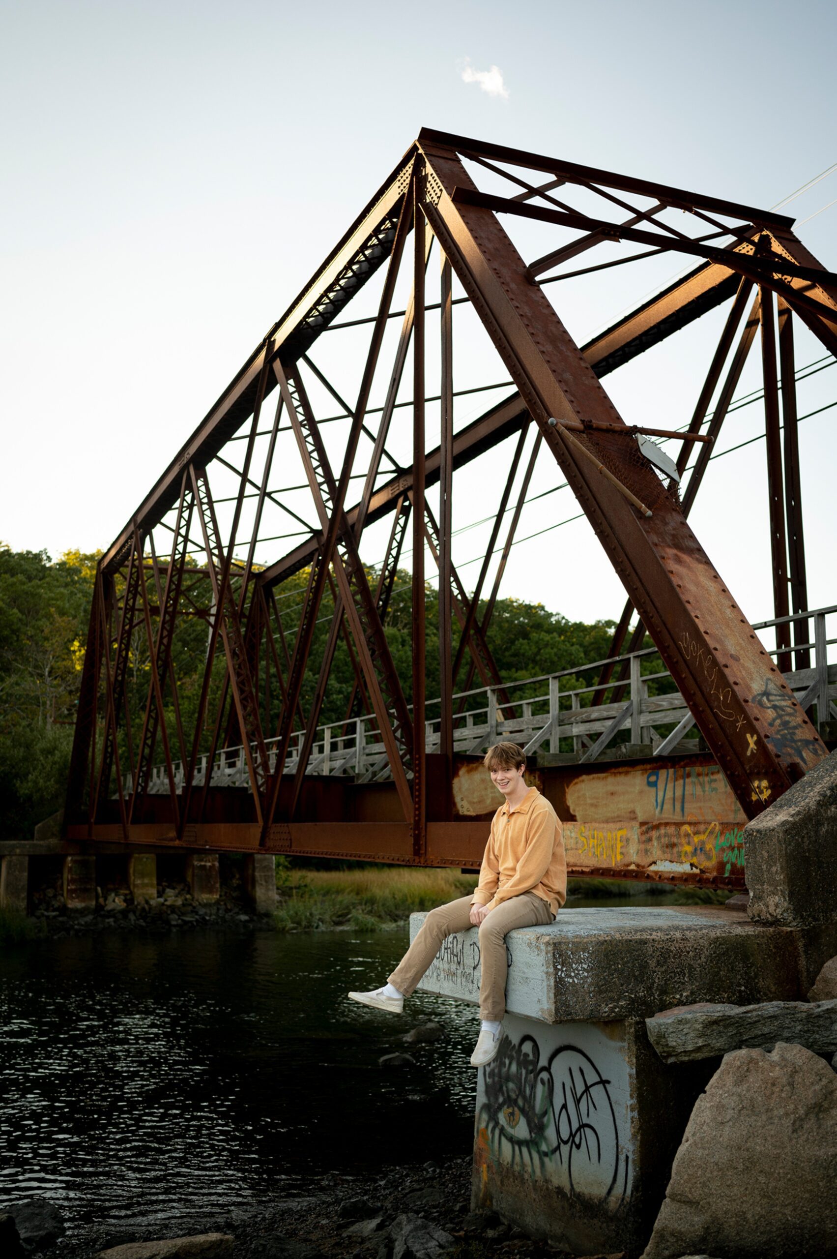 A senior sits on a bridge support over a river in an orange shirt and pants after attending private high schools in connecticut