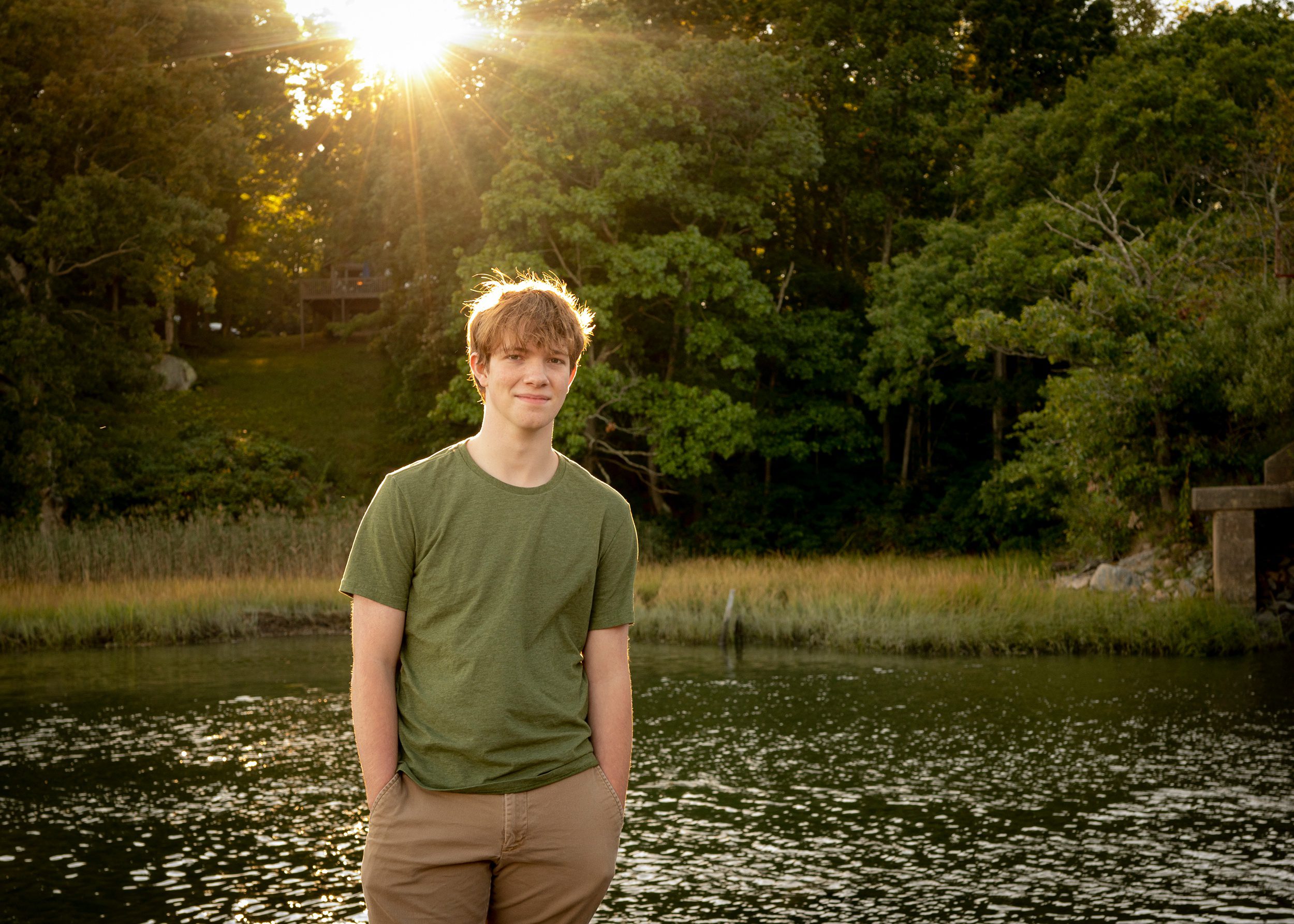 A senior from private high schools in connecticut stands by a pond at sunset in a green shirt with hands in pockets
