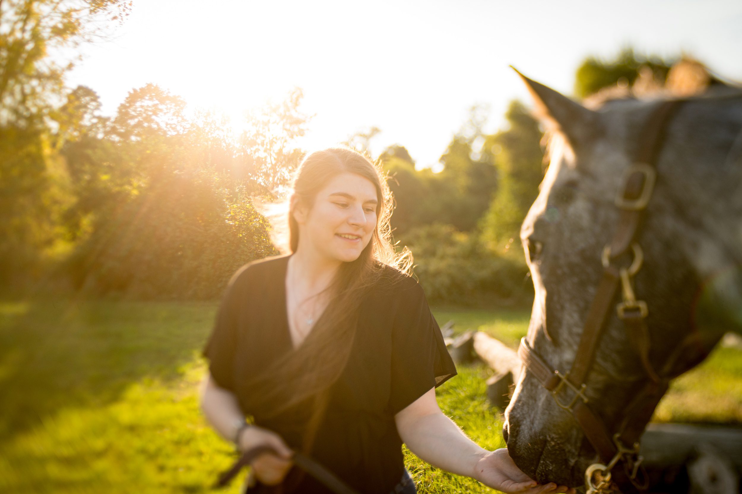 A high school senior feeds a grey horse in a pasture at sunset in a black dress after attending sacred heart academy hamden
