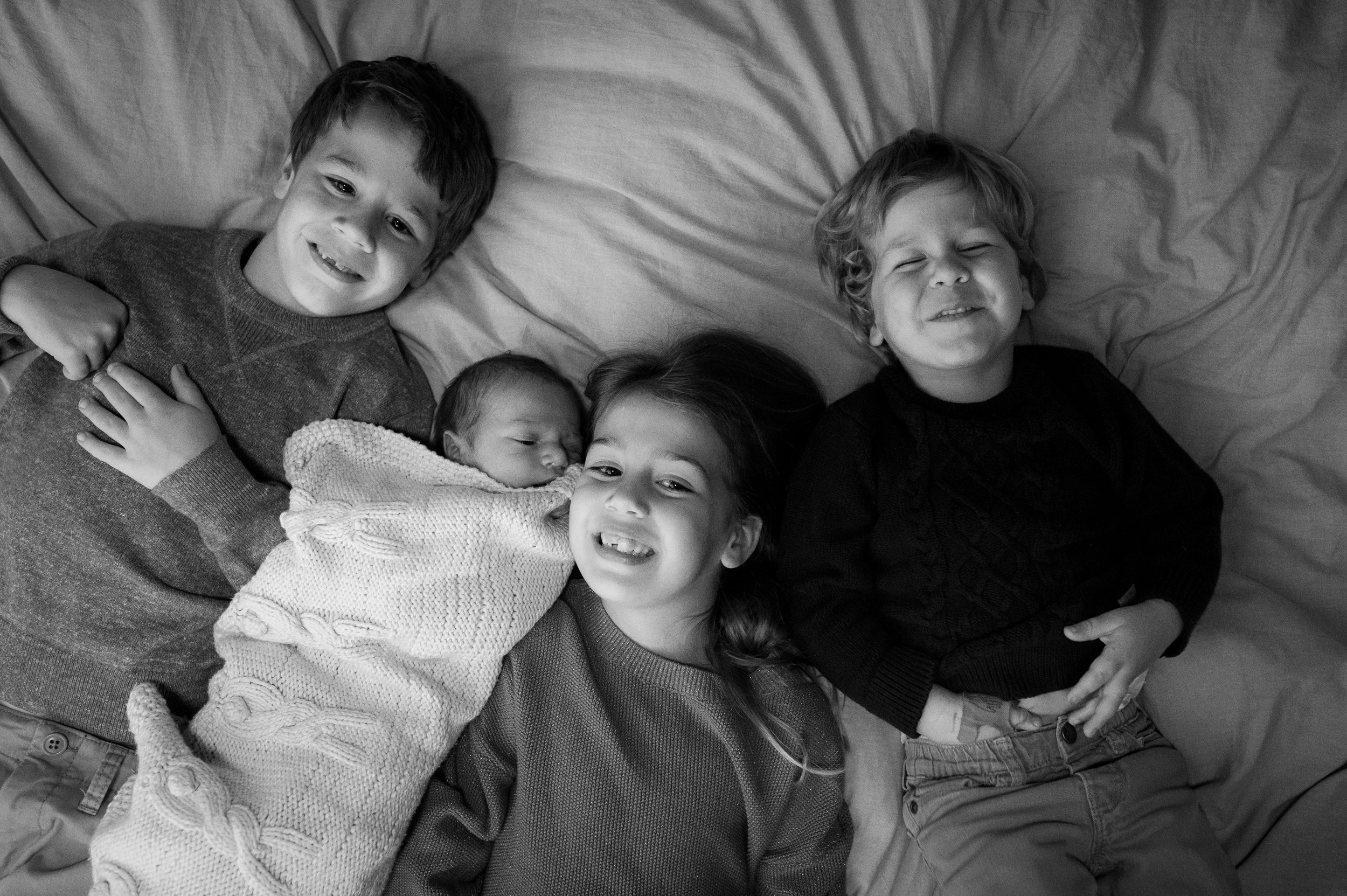 Three young siblings lay on a bed snuggling their sleeping newborn sibling and smiling during things to do in new haven with kids