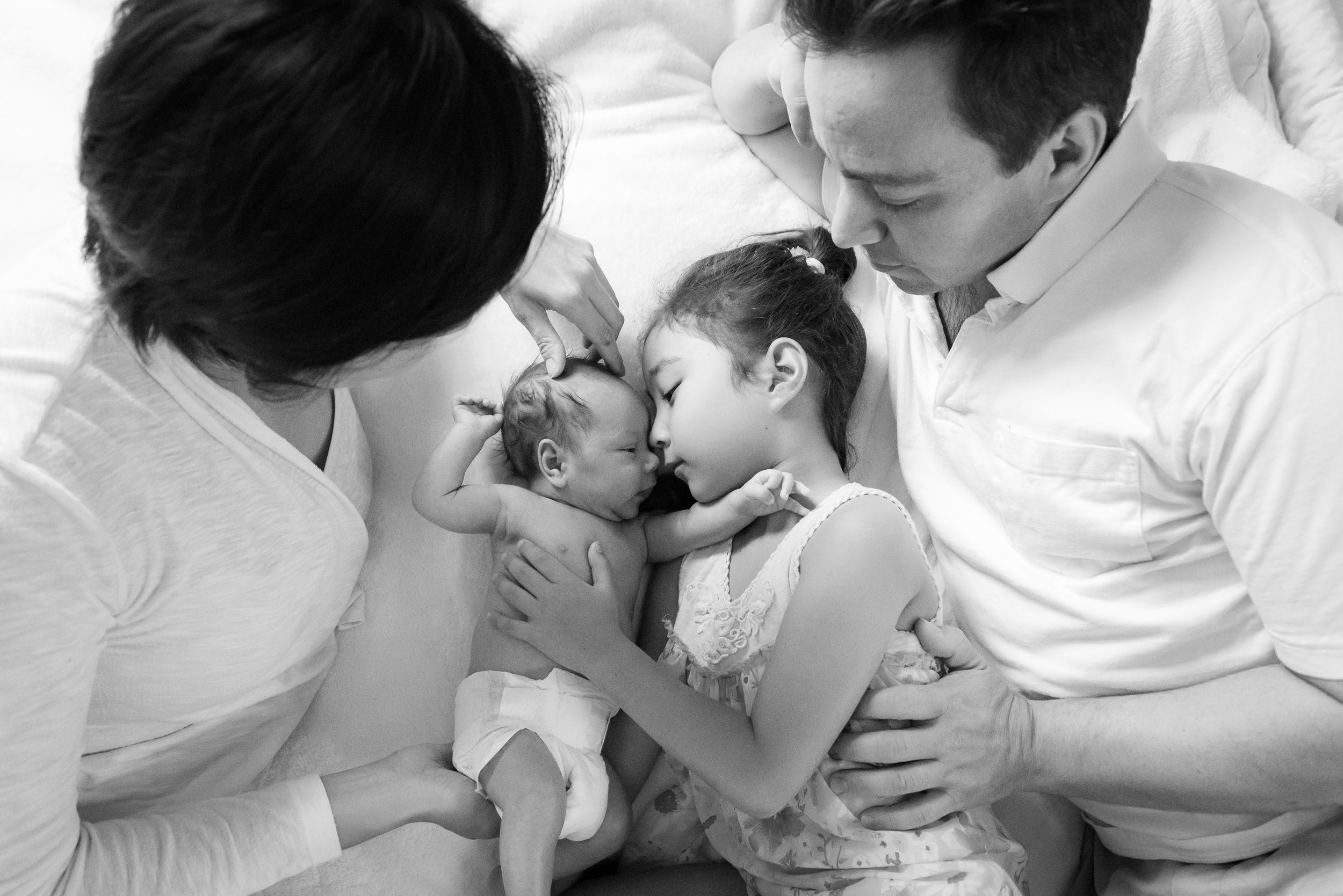 A toddler girl snuggles with her newborn baby sibling and parents on a bed