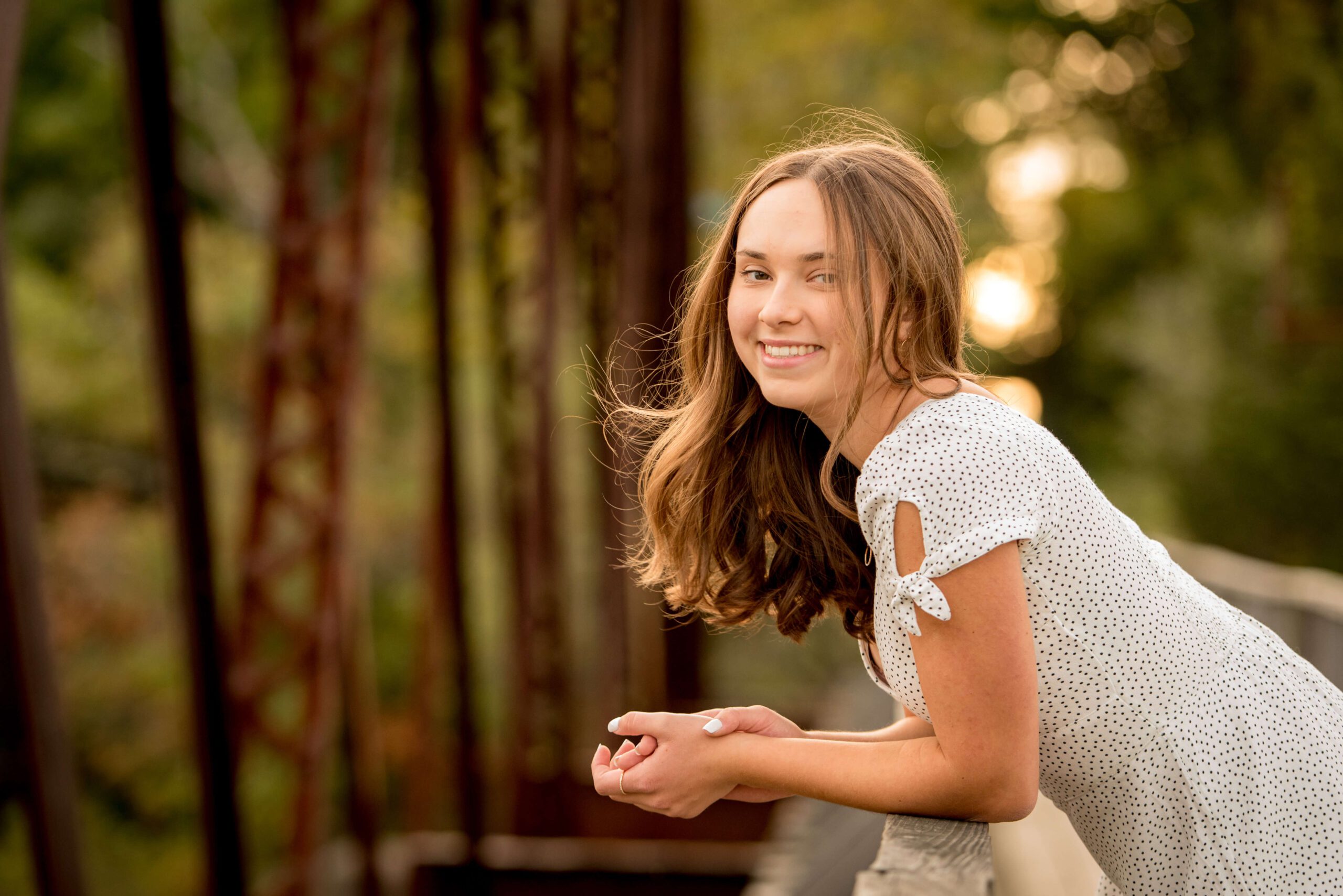A teenage girl leans on a wooden bridge railing at sunset while smiling