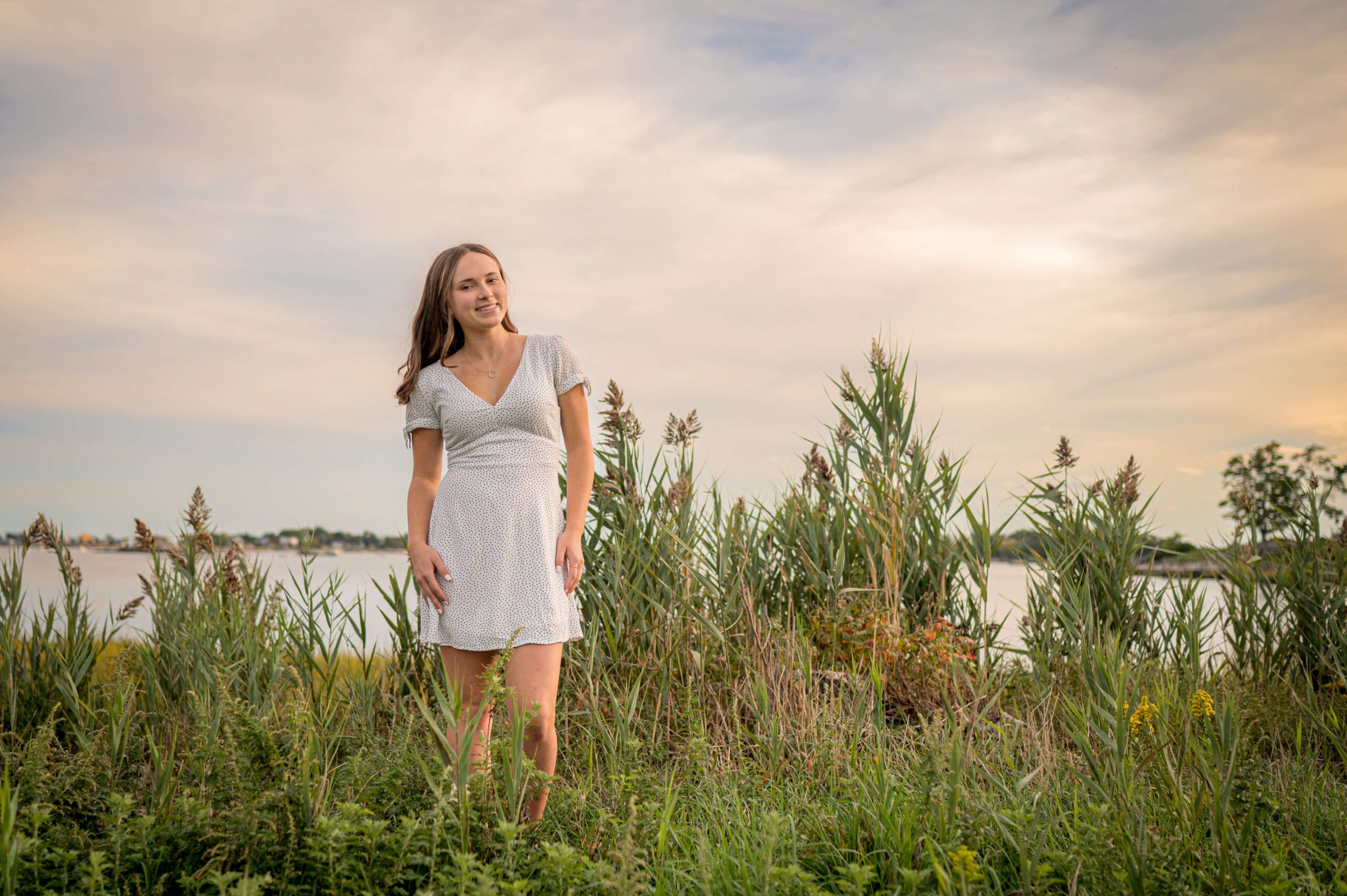 A high school senior in a white dres walks in tall grasses by the water with a smiling after visiting nail salons in madison ct