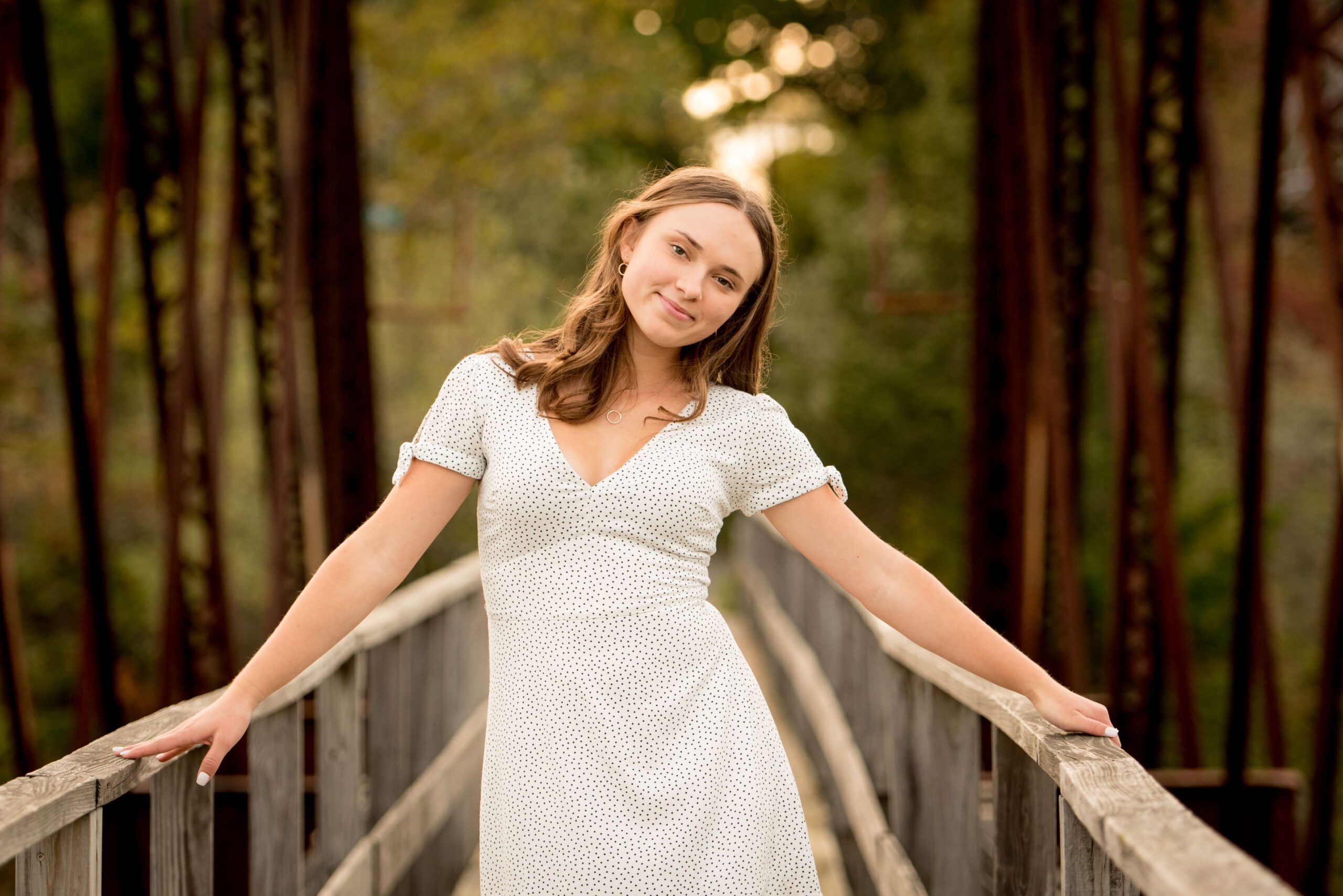 A high school senior in a white dress stands with matching white nails on a wooden bridge at sunset with a smile after visiting nail salons in madison ct