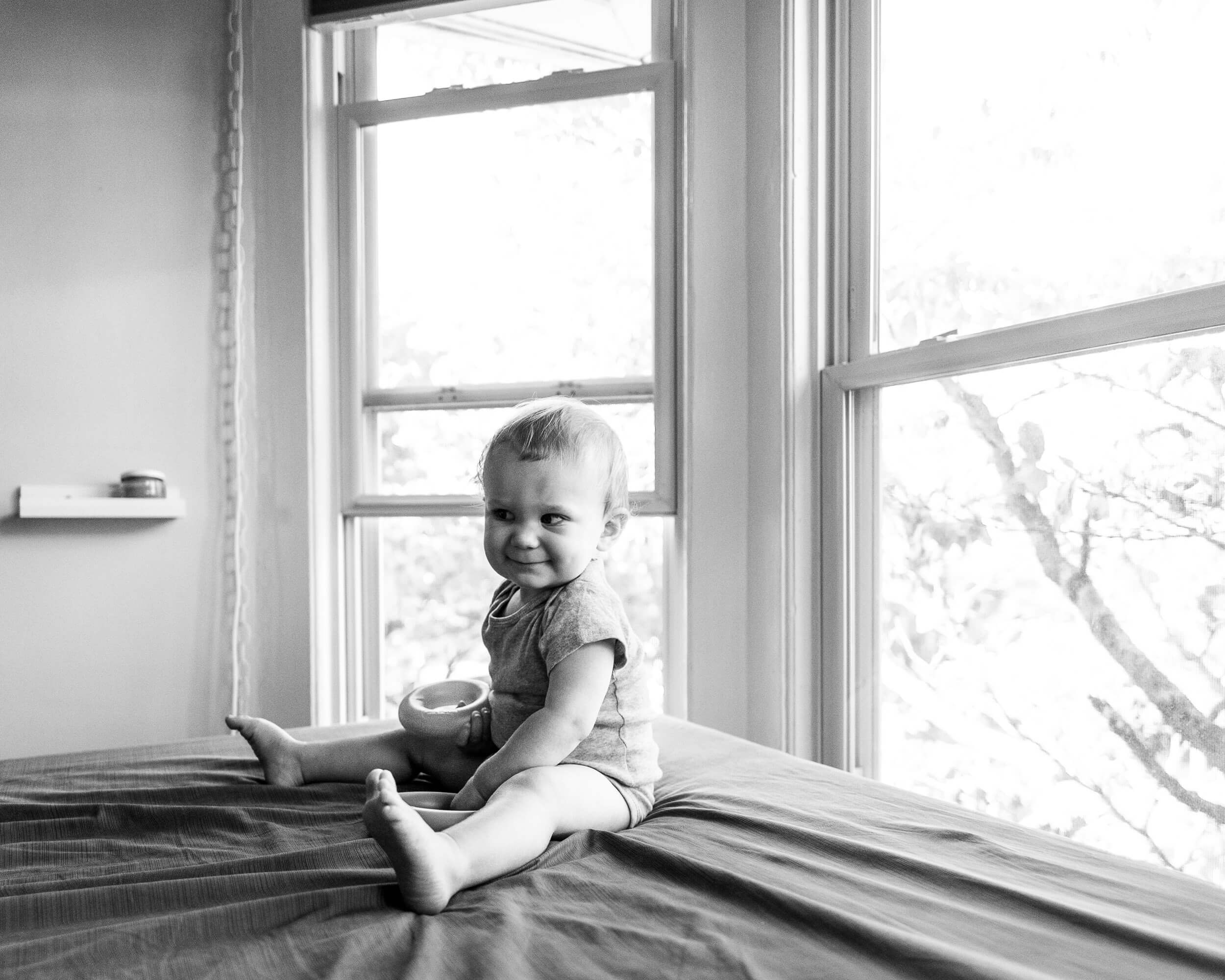 a toddler girl in a onesie sits on a bed under a window with a smile and a ring toy in black and white after visiting pediatric dentists in connecticut