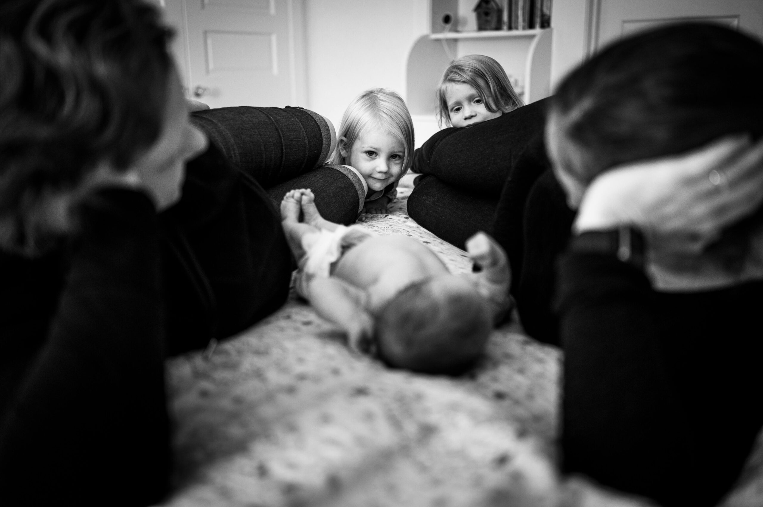 A newborn baby lays between parents on a bed with toddler big sisters peeking in after meeting lactation consultants in CT