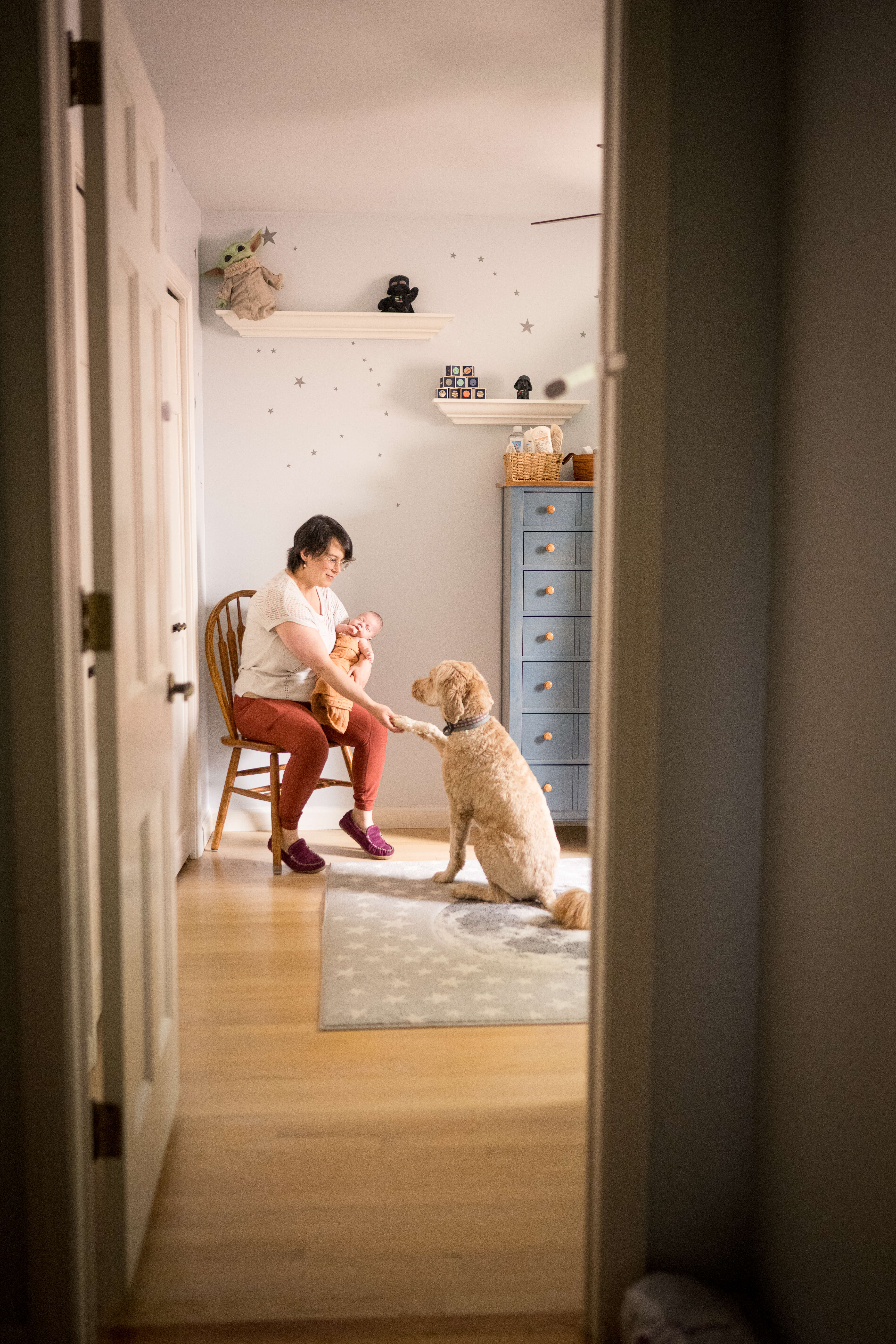 A woman sits in a kitchen chair holding a sleeping newborn while shaking paws with a golden doodle after some prenatal yoga in connecticut