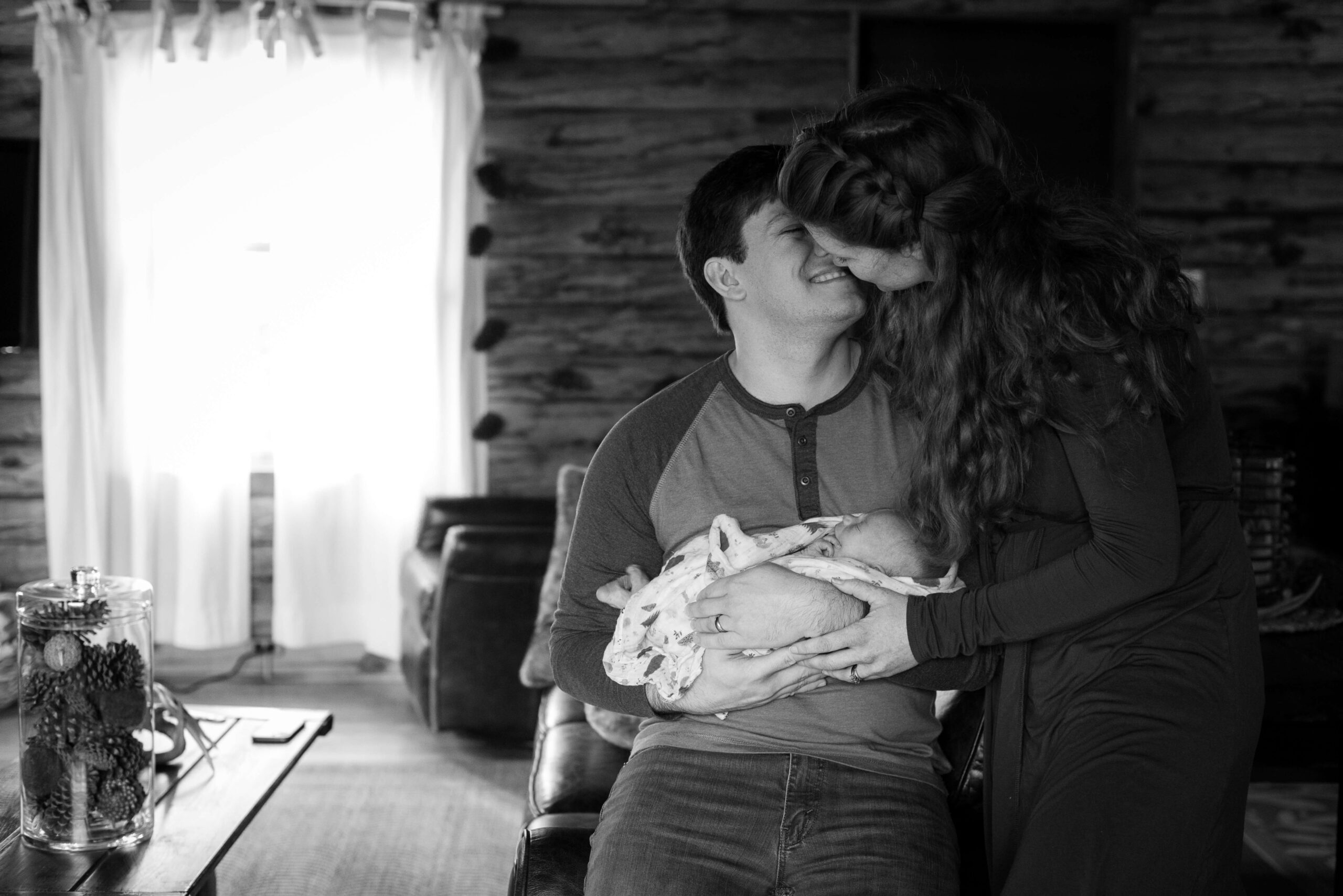 New parents lean for a kiss while dad cradles their sleeping newborn and leans on a couch after visiting flutterby in guilford