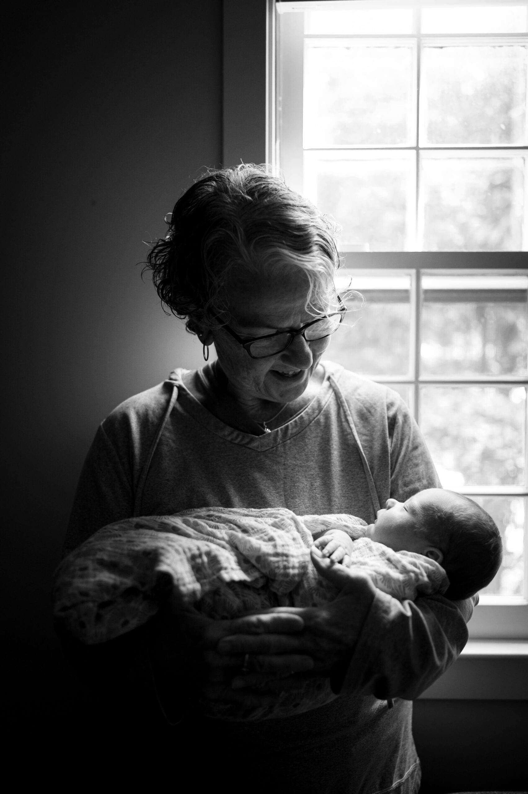 A happy grandma stands in a window smiling down at her sleeping newborn grandbaby in her arms after visiting flutterby in guilford