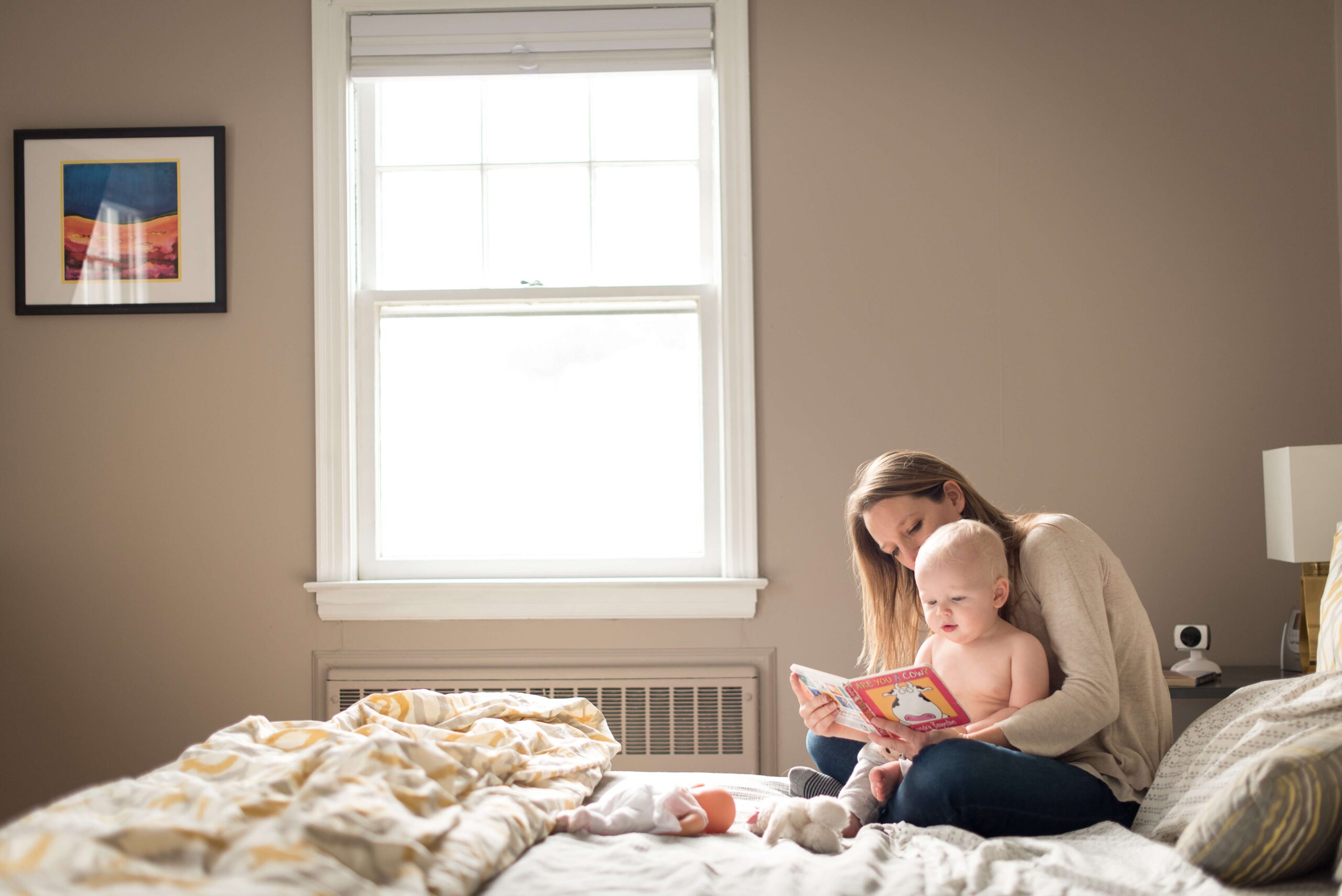 A mother in a tan sweater read a book to her baby in her lap on a bed under a window after using great obgyns in connecticut