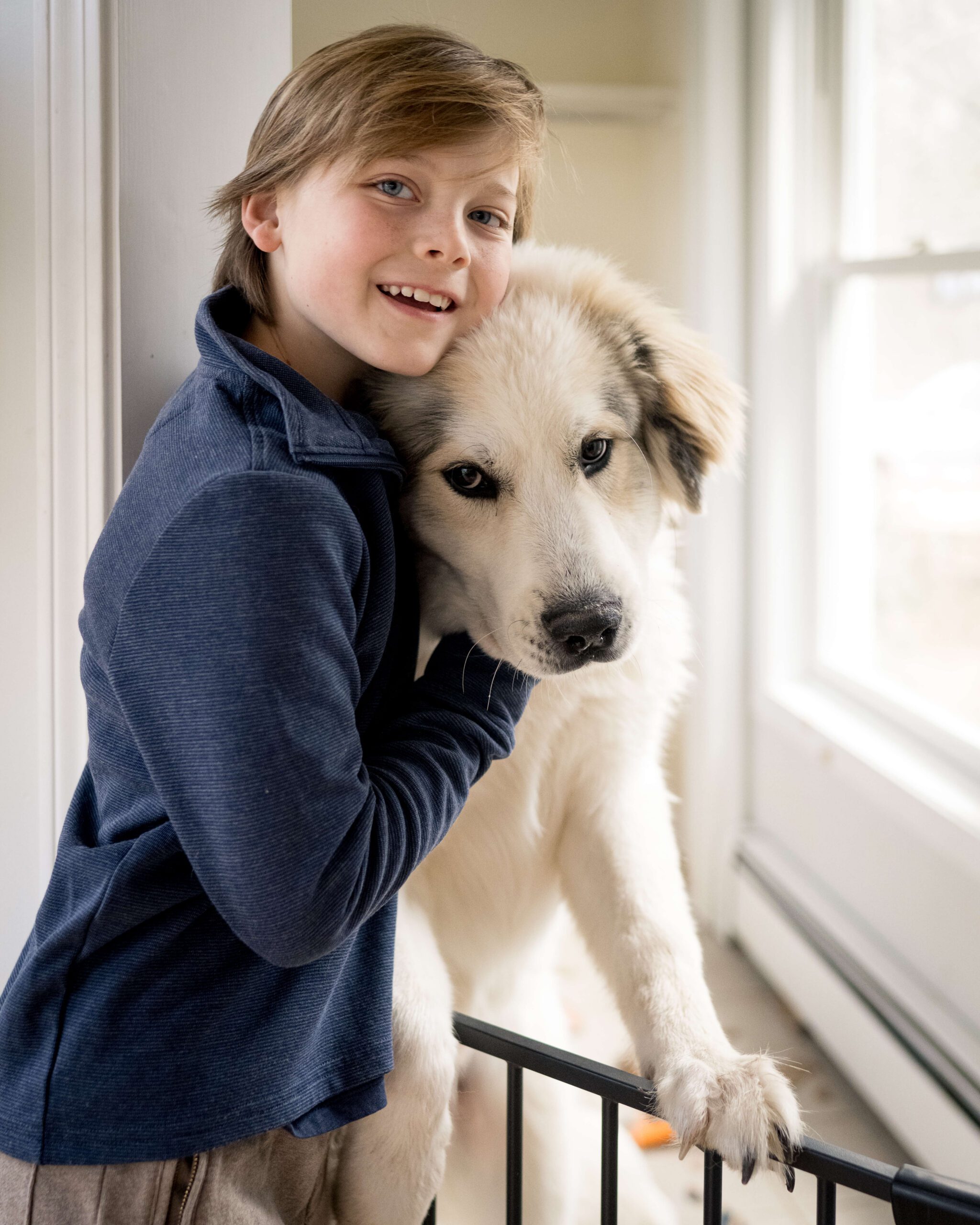 A young boy hugs a big white dog while standing in a hall in a blue shirt